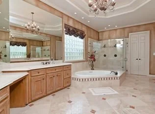 Spacious bathroom with a large mirror no countertop, wooden cabinets, a glass-enclosed shower, and a bathtub with a floral arrangement on the side, illuminated by chandeliers and natural light from multiple windows.