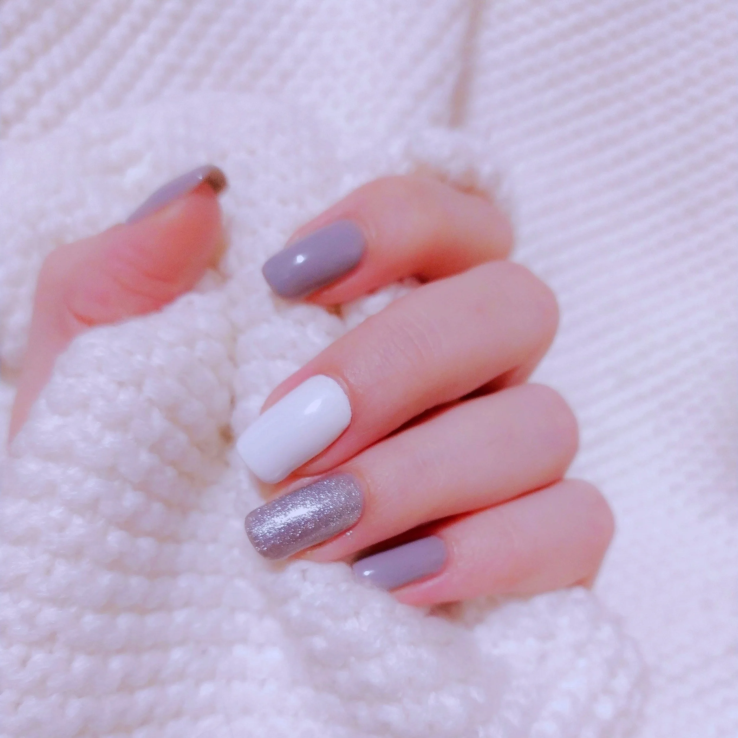 Close-up of a hand with neatly manicured nails painted in various shades of white, gray, and glitter, resting on a soft, textured white surface.