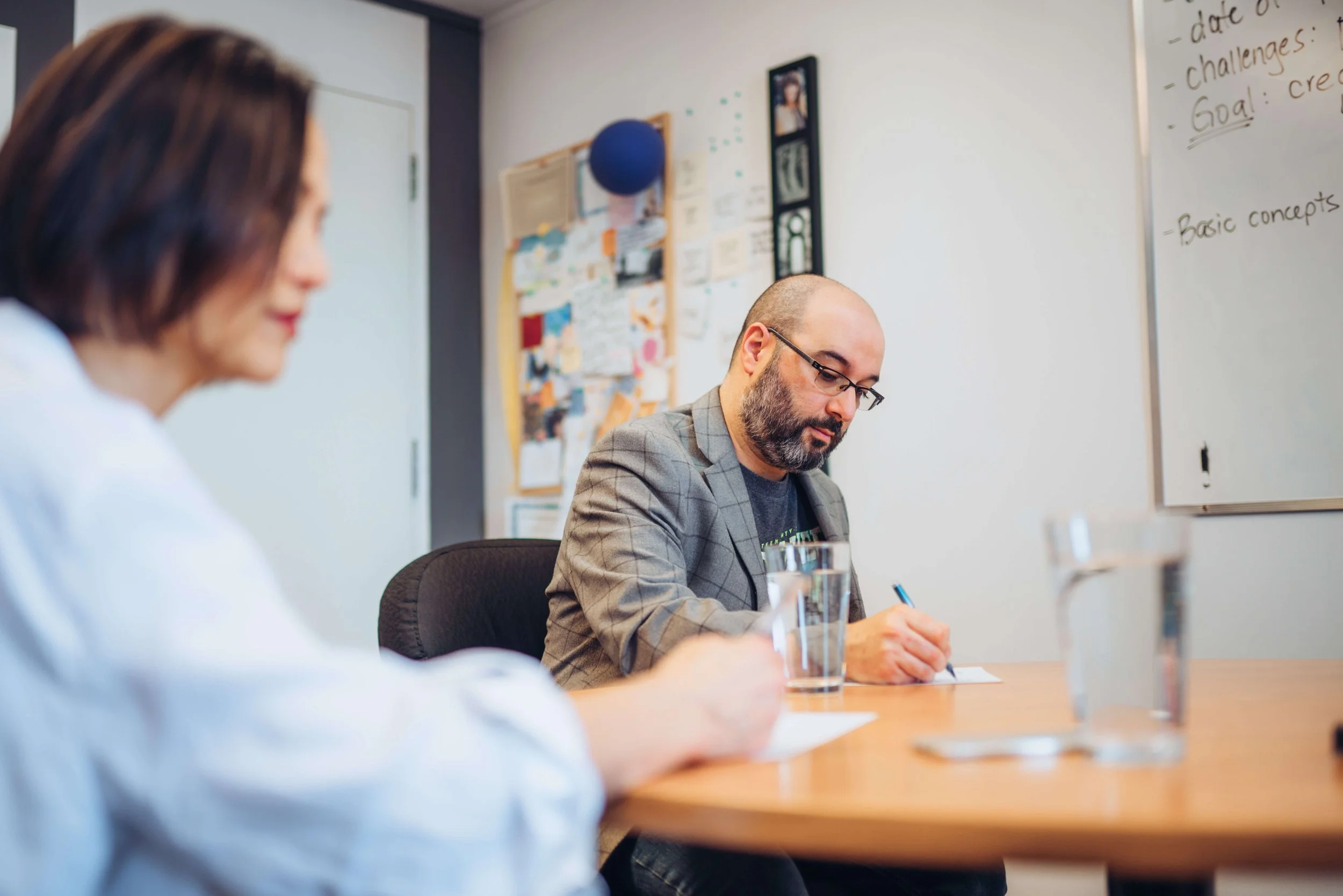 Two people are sitting at a conference table in a meeting room with a whiteboard and bulletin board behind them. One person, a woman with shoulder-length brown hair, is blurred in the foreground. The other person, a bald man with glasses and a beard, is wearing a gray blazer and is focused on writing on a piece of paper. There are glasses of water on the table.