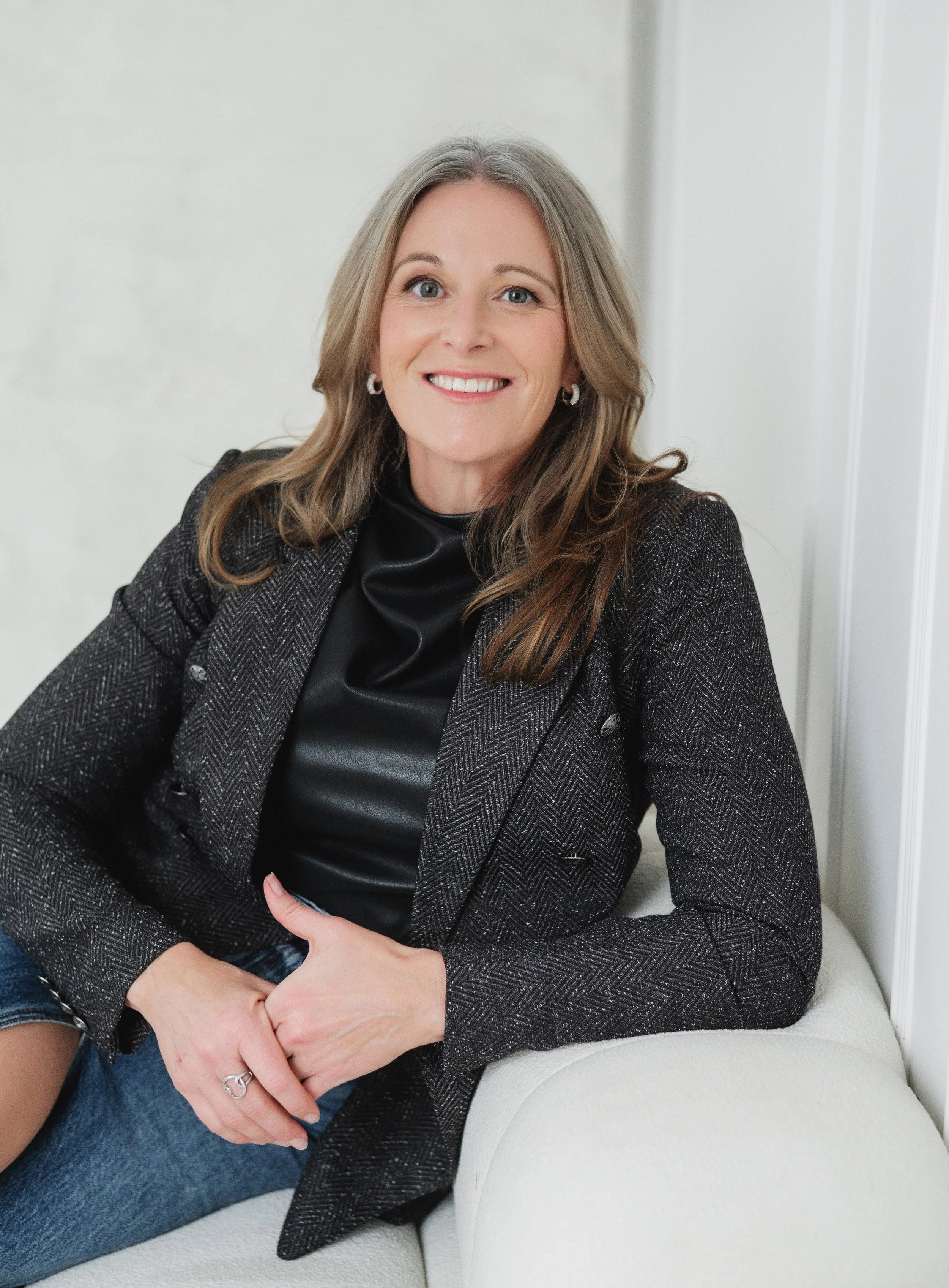 Separation and divorce coach Lana Wickstrom in Saskatoon is wearing a black top and a gray blazer, sitting on a white couch with a light-colored wall in the background.
