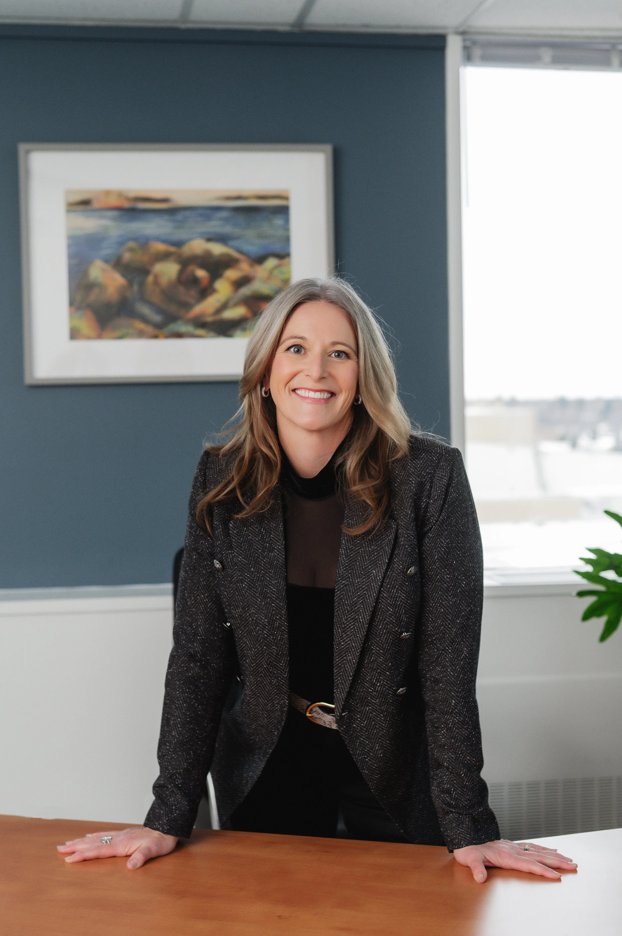 Separation and divorce coach Lana Wickstrom in Saskatoon is leaning on her desk, with a painted landscape on the dark blue wall behind her and a window with bright light to the right.