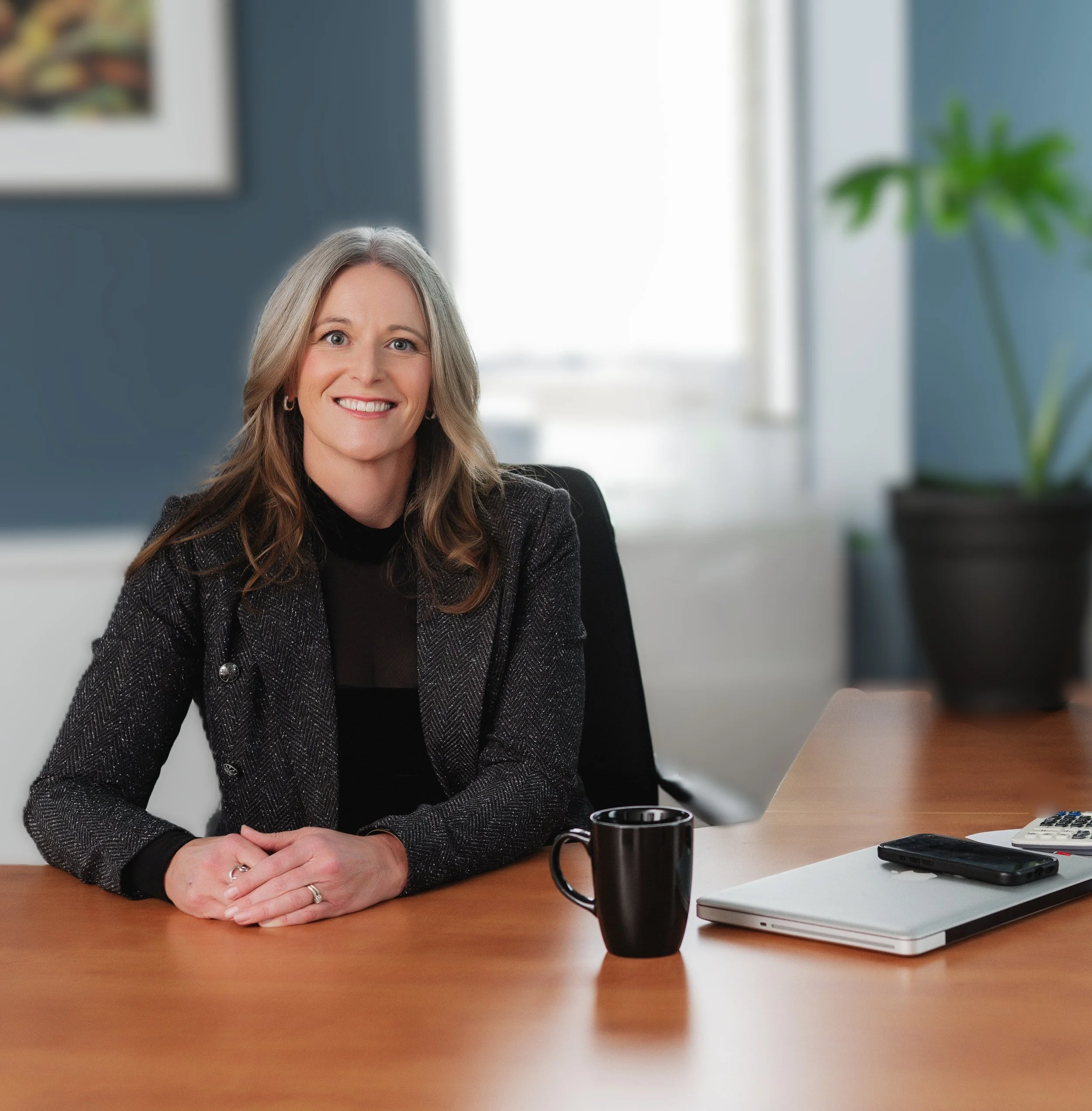 Separation and divorce coach Lana Wickstrom in Saskatoon is sitting at her desk in her office. The background shows a bright window, a blue wall, a framed picture, and a green potted plant.