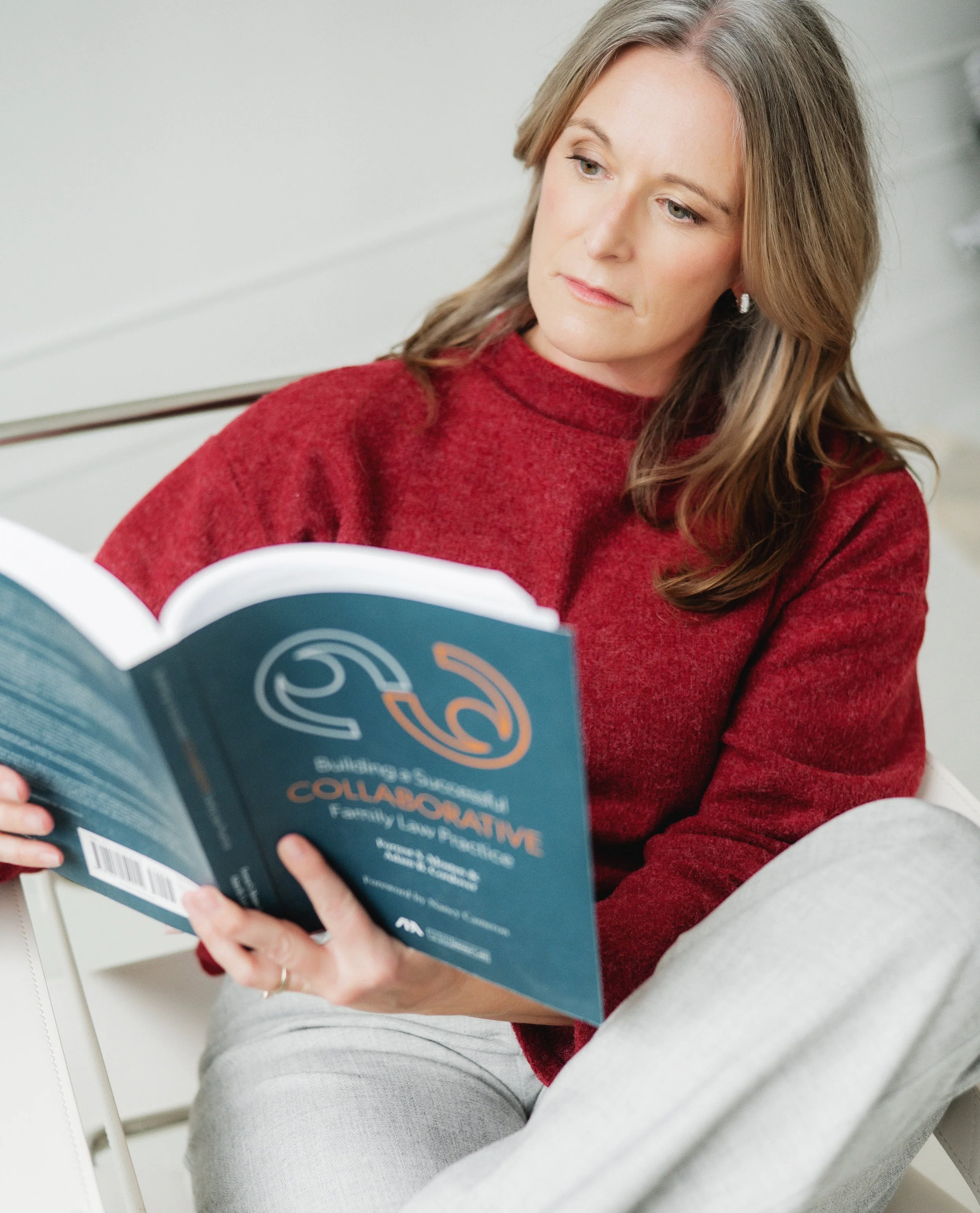 Separation and divorce coach Lana Wickstrom in Saskatoon is wearing a red sweater reading a book titled 'Collaborative Family Law Practice' in a well-lit room.