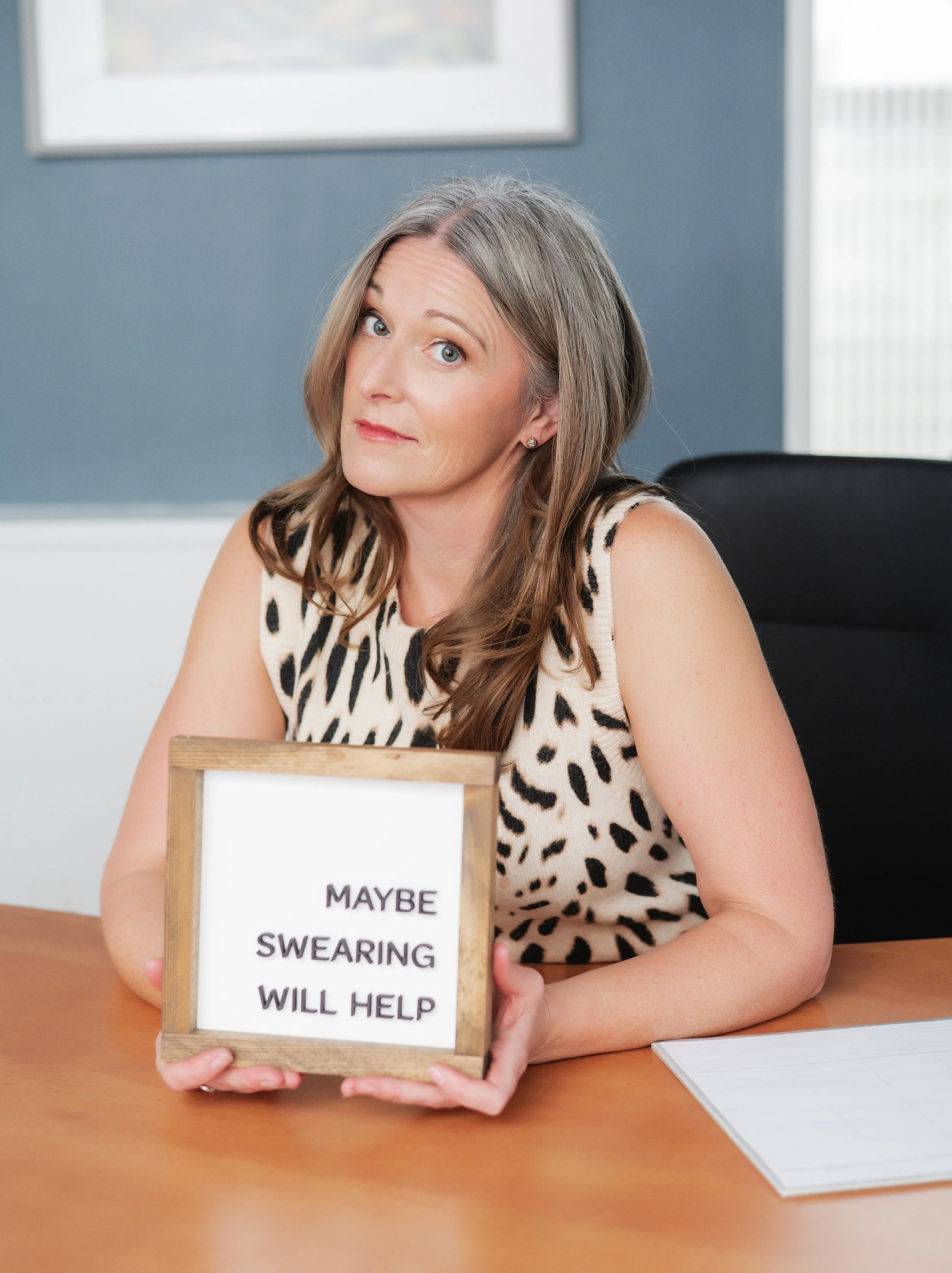 Separation and divorce coach Lana Wickstrom in Saskatoon is sitting at a wooden table, holding a wooden framed sign that says, "MAYBE SWEARING WILL HELP."