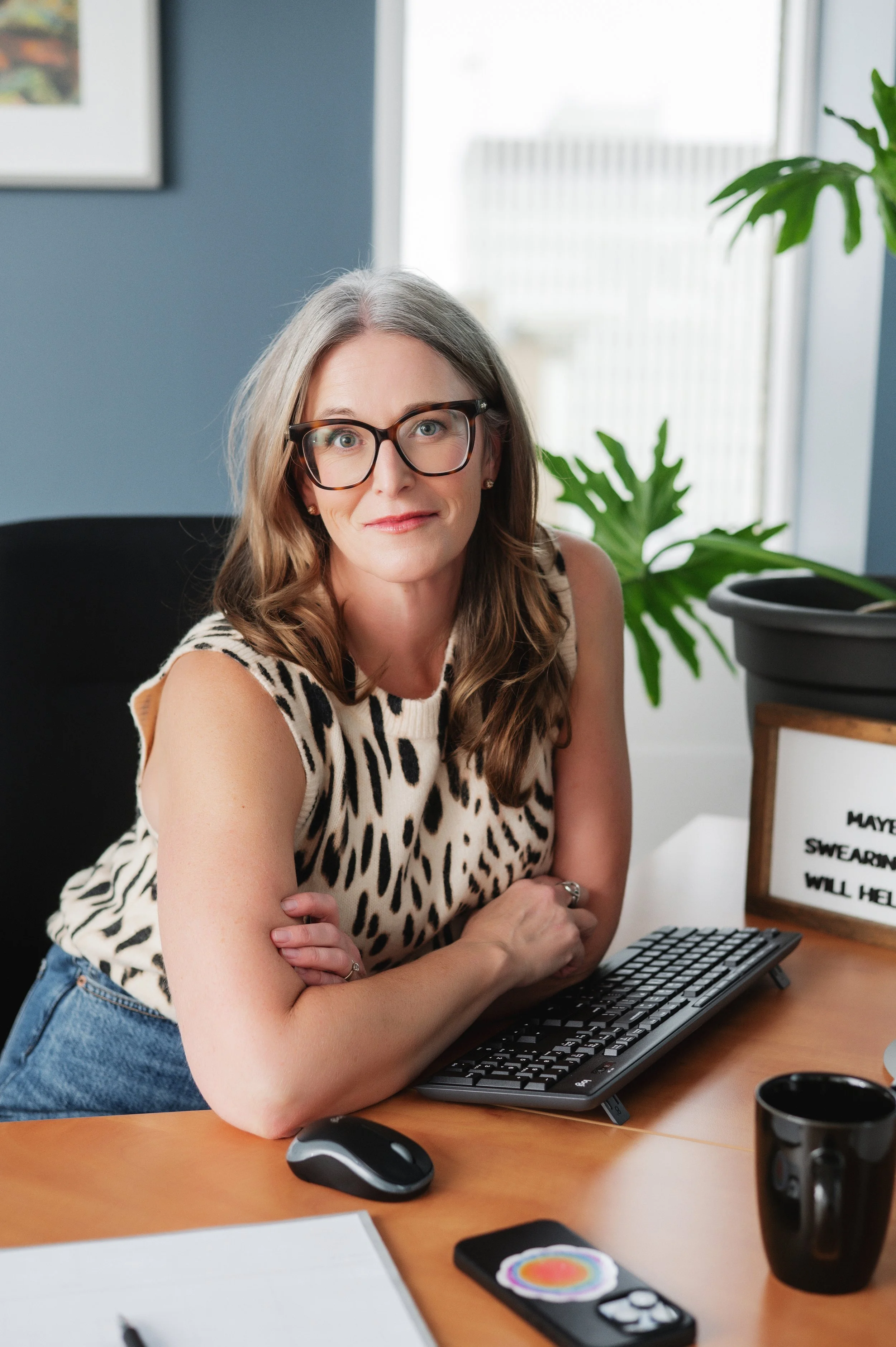 A woman with glasses and long hair sitting at a desk with her arms crossed, using a computer keyboard, with a monitor and various office supplies on the desk, in a room with a blue wall, framed artwork, and a window with natural light.