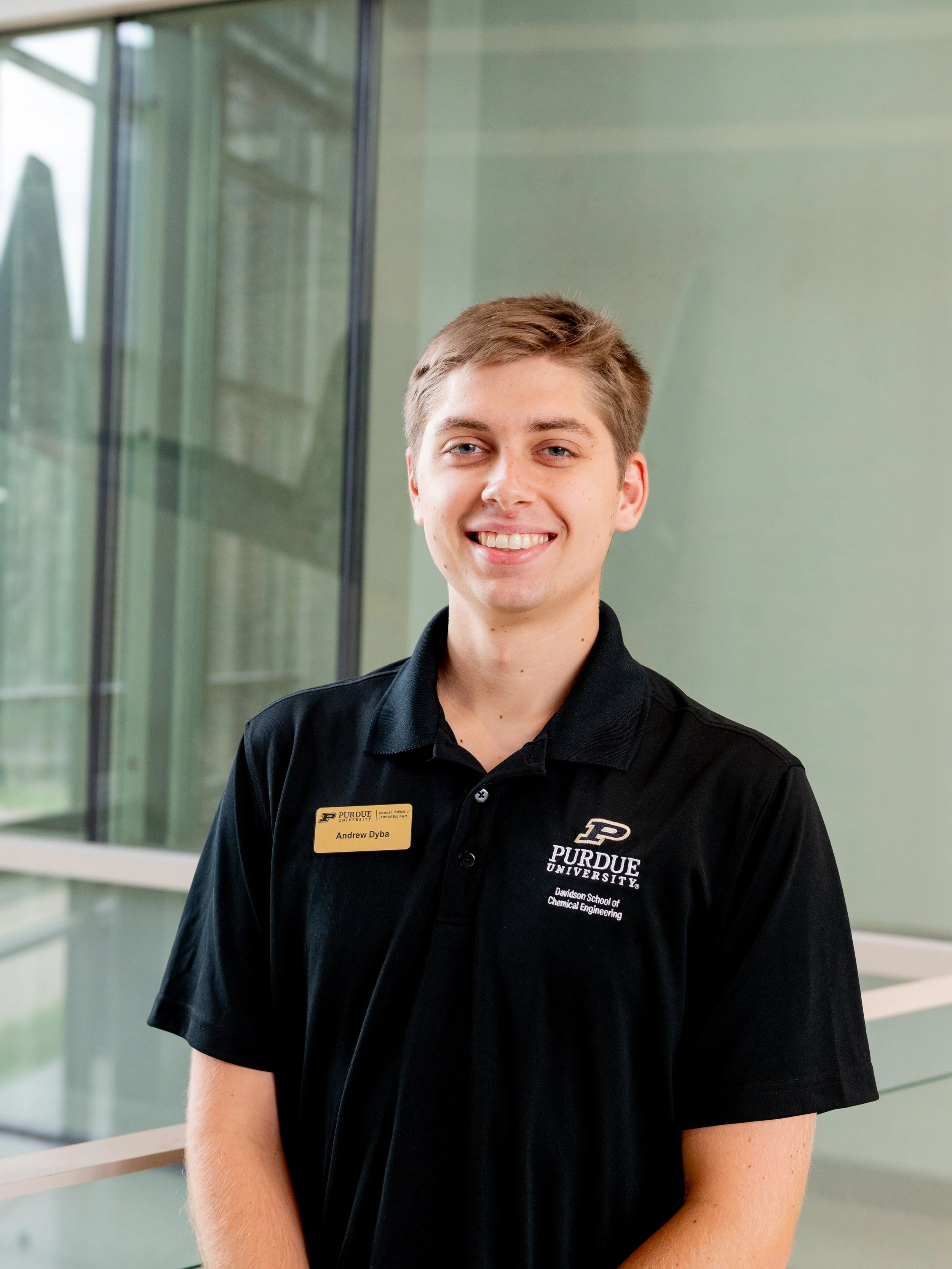 A young woman with black hair and a black Purdue University polo shirt, standing indoors in front of a brick wall and windows, smiling at the camera.