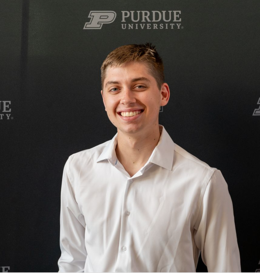 A young woman with black hair and a black Purdue University polo shirt, standing indoors in front of a brick wall and windows, smiling at the camera.