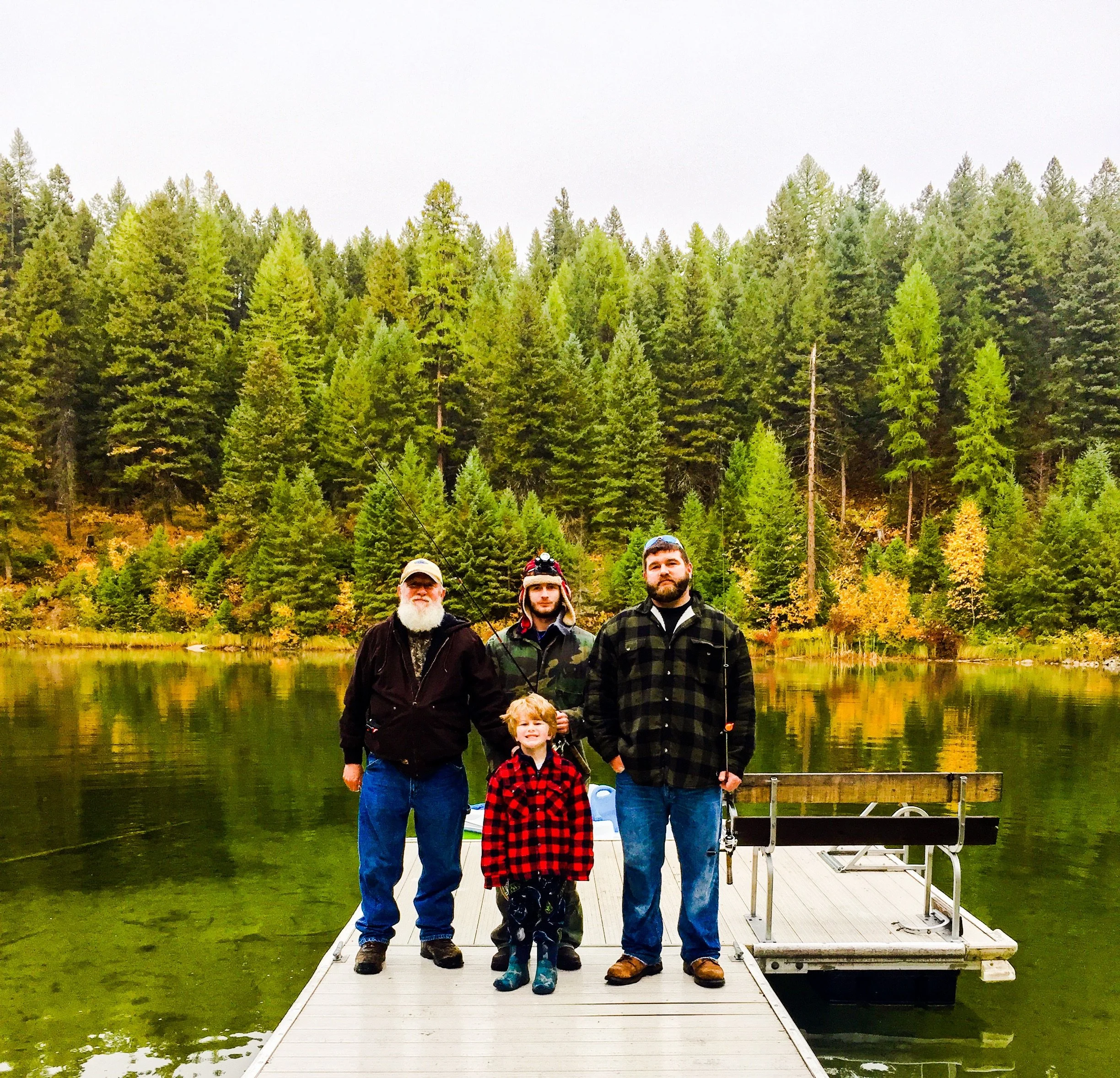 Four people, an adult man, a young man, an elderly man, and a young boy, standing on a dock by a lake, with a forested area in the background. The scene suggests they are enjoying a fishing trip in Montana.