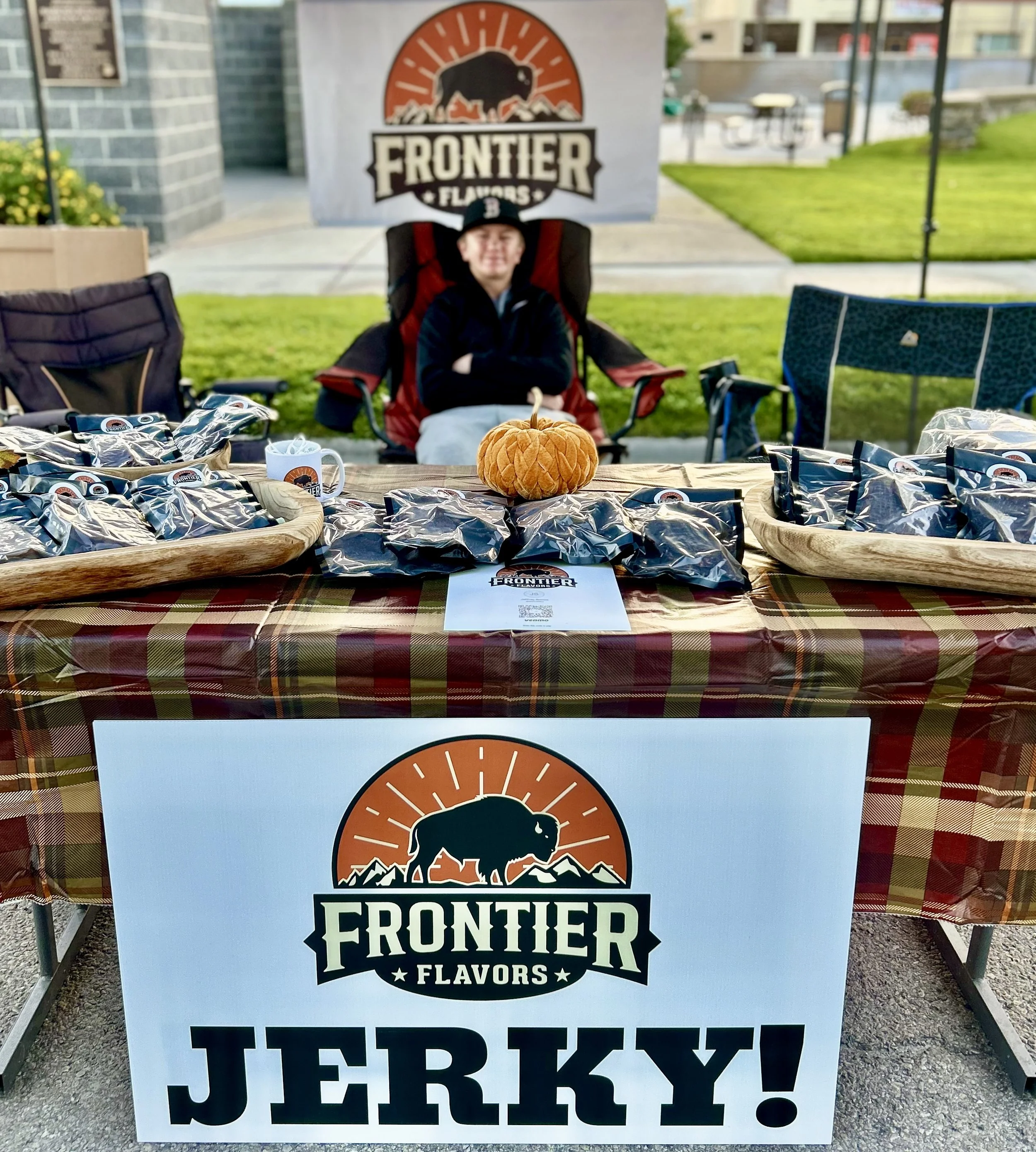 A boy sitting at an outdoor booth for Frontier Flavors, with a banner behind him and a sign in front that says "JERKY!" The booth has a pumpkin and packages of Ribeye Style jerky on the table.