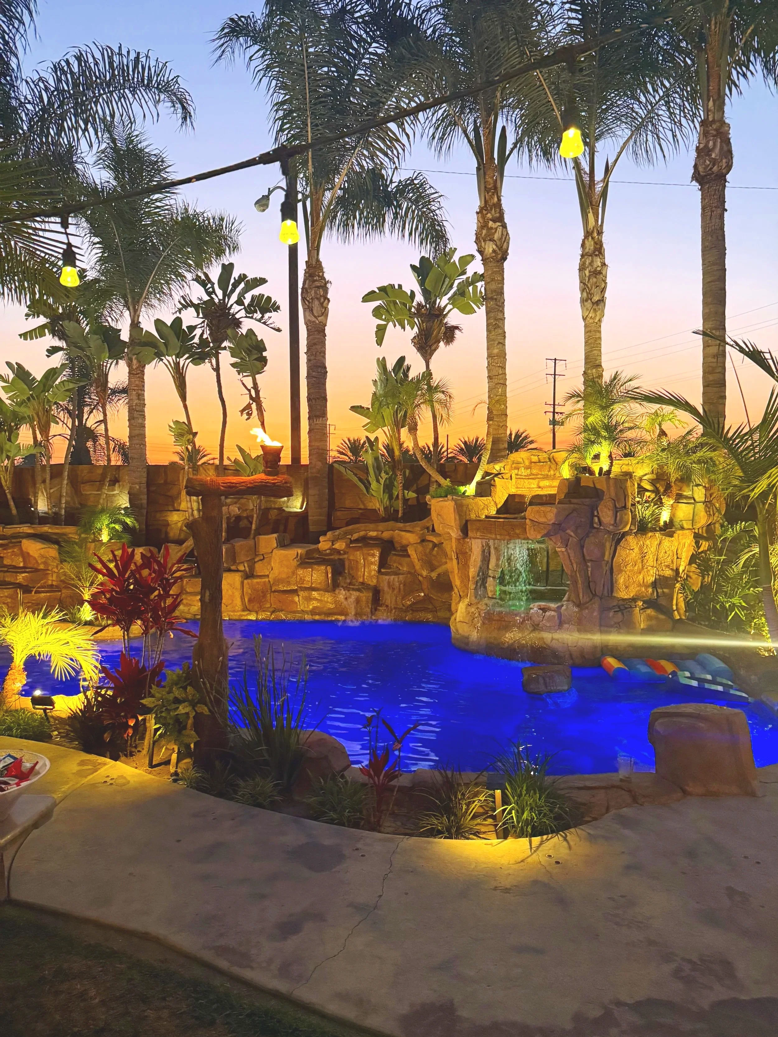 A backyard with a swimming pool illuminated with blue and yellow lights, surrounded by palm trees and tropical plants, at sunset with a colorful sky.
