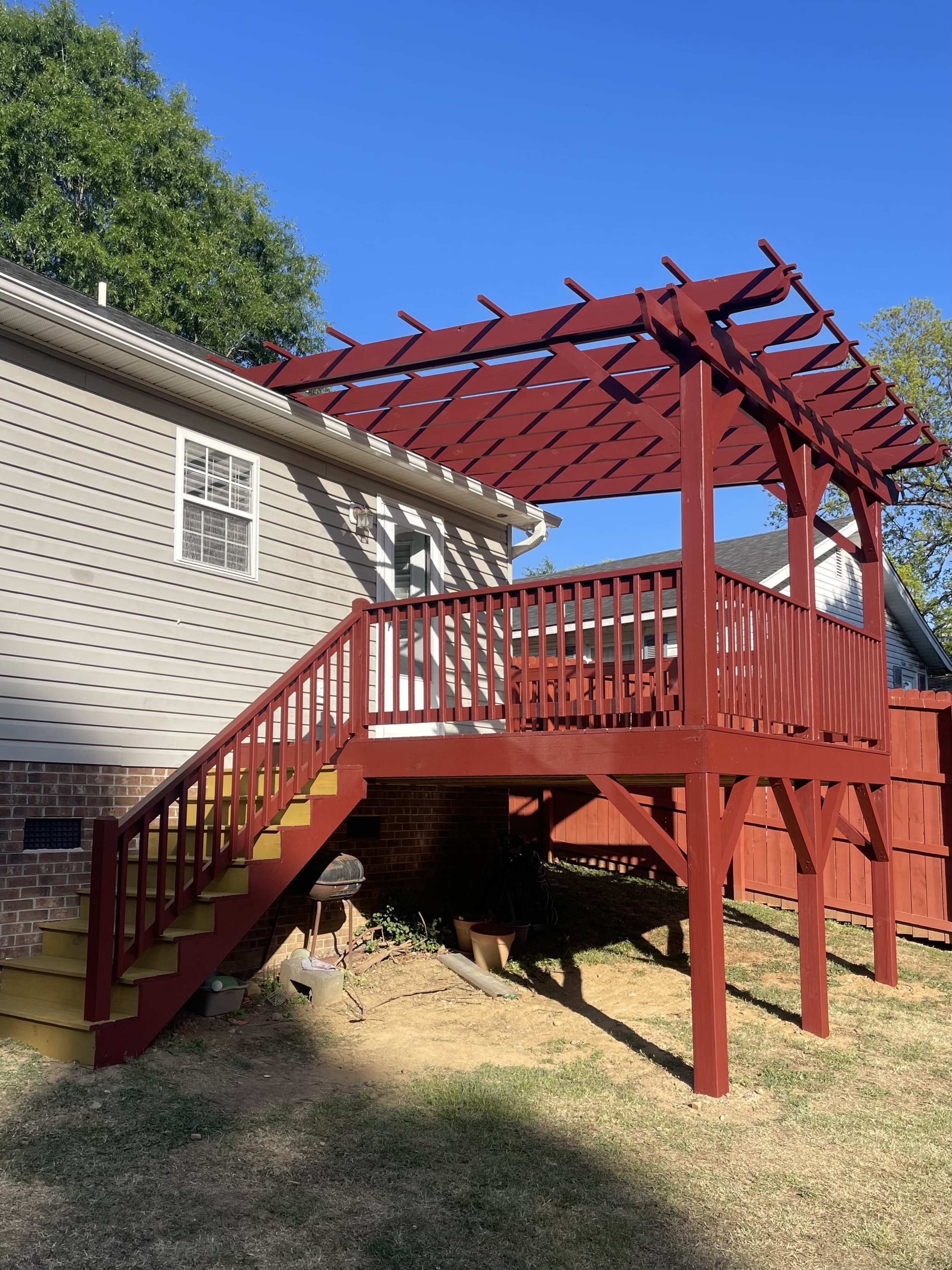 Backyard view featuring a beige house with an upstairs window, a red wooden deck with stairs, a barbecue grill, empty flower pots, and a fenced yard with green grass and trees under a clear blue sky.