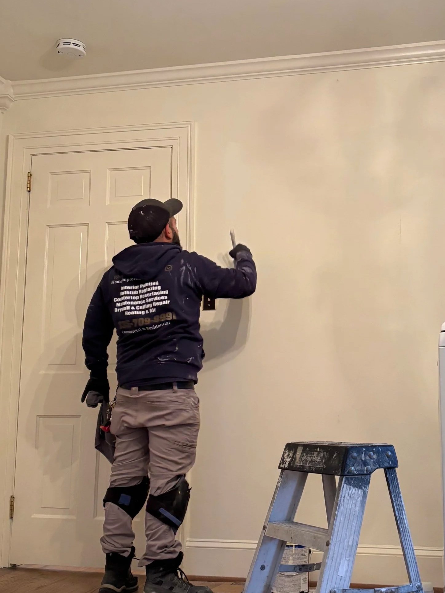 A worker in protective gear painting a wall with a paintbrush. He is standing on the floor near a step ladder with paint supplies. The wall is cream-colored, and there is a closed door to his left.