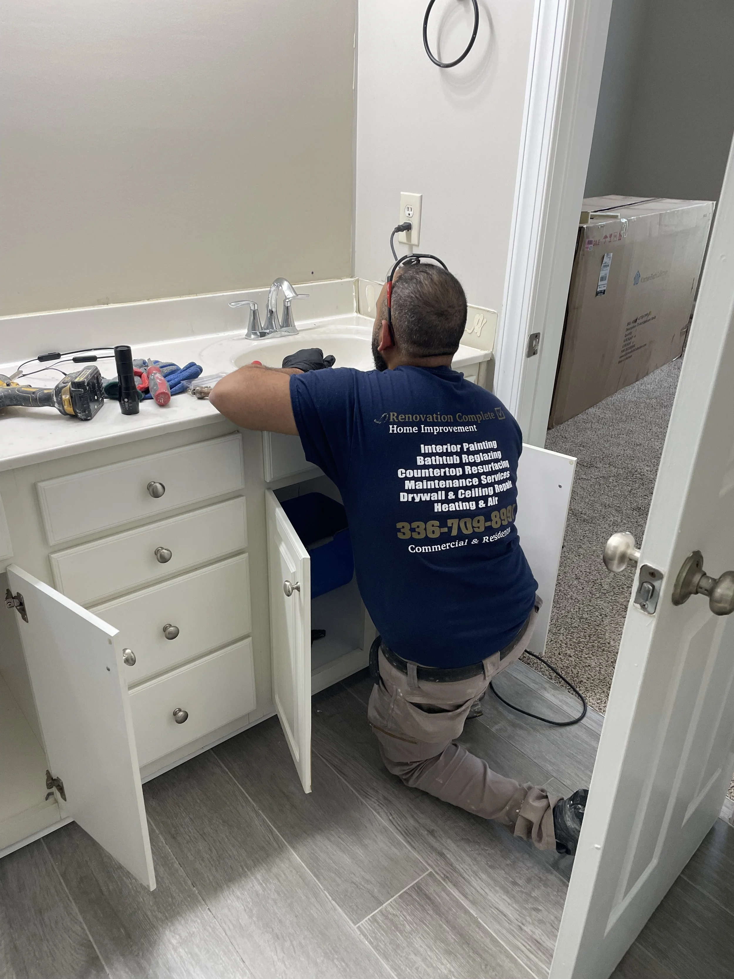 A handyman kneeling on the floor working on the plumbing of a bathroom vanity cabinet. The cabinet door is open, and tools are on the countertop.