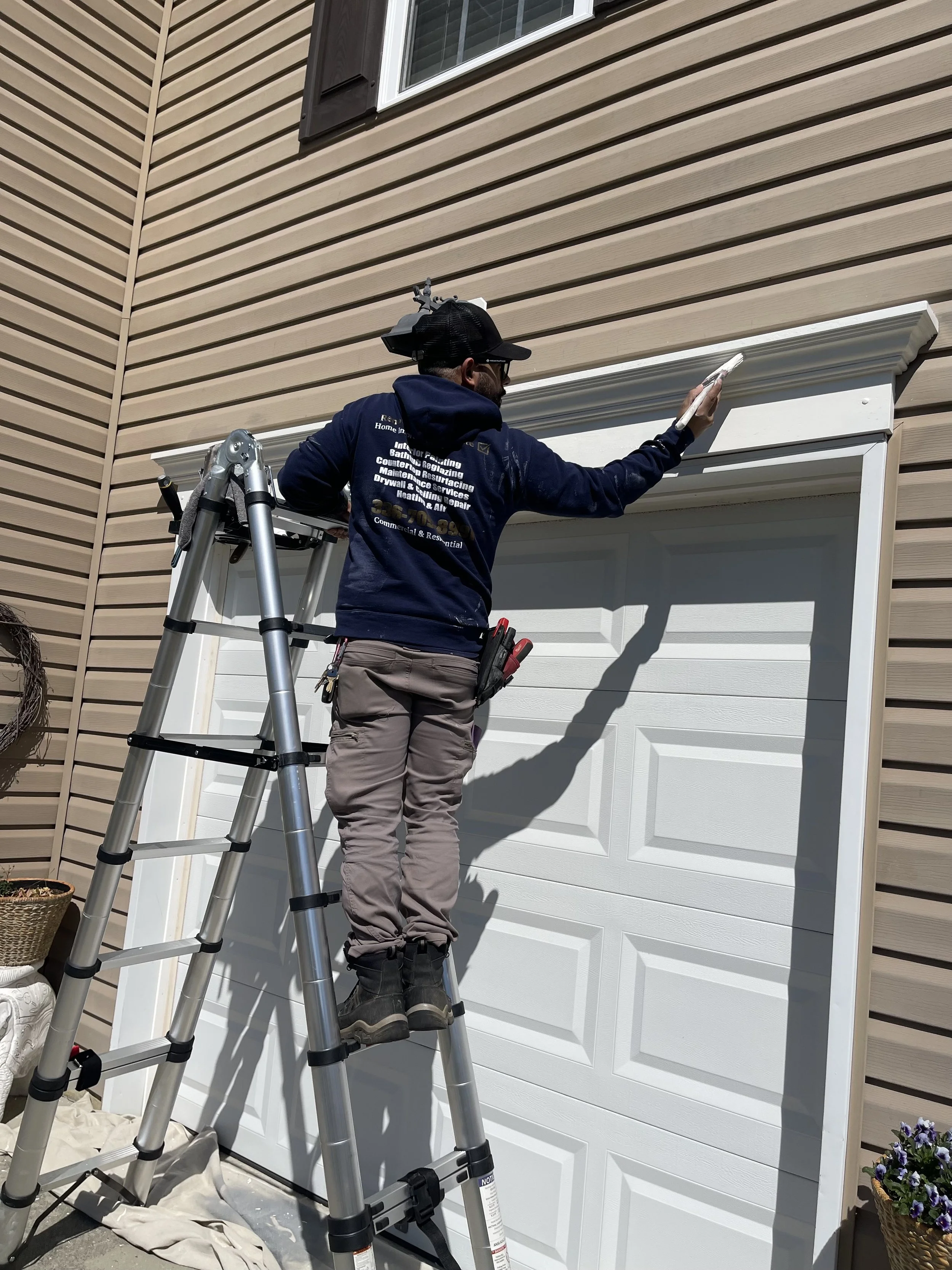 A man on a ladder painting trim above a garage door outside a house with beige siding.