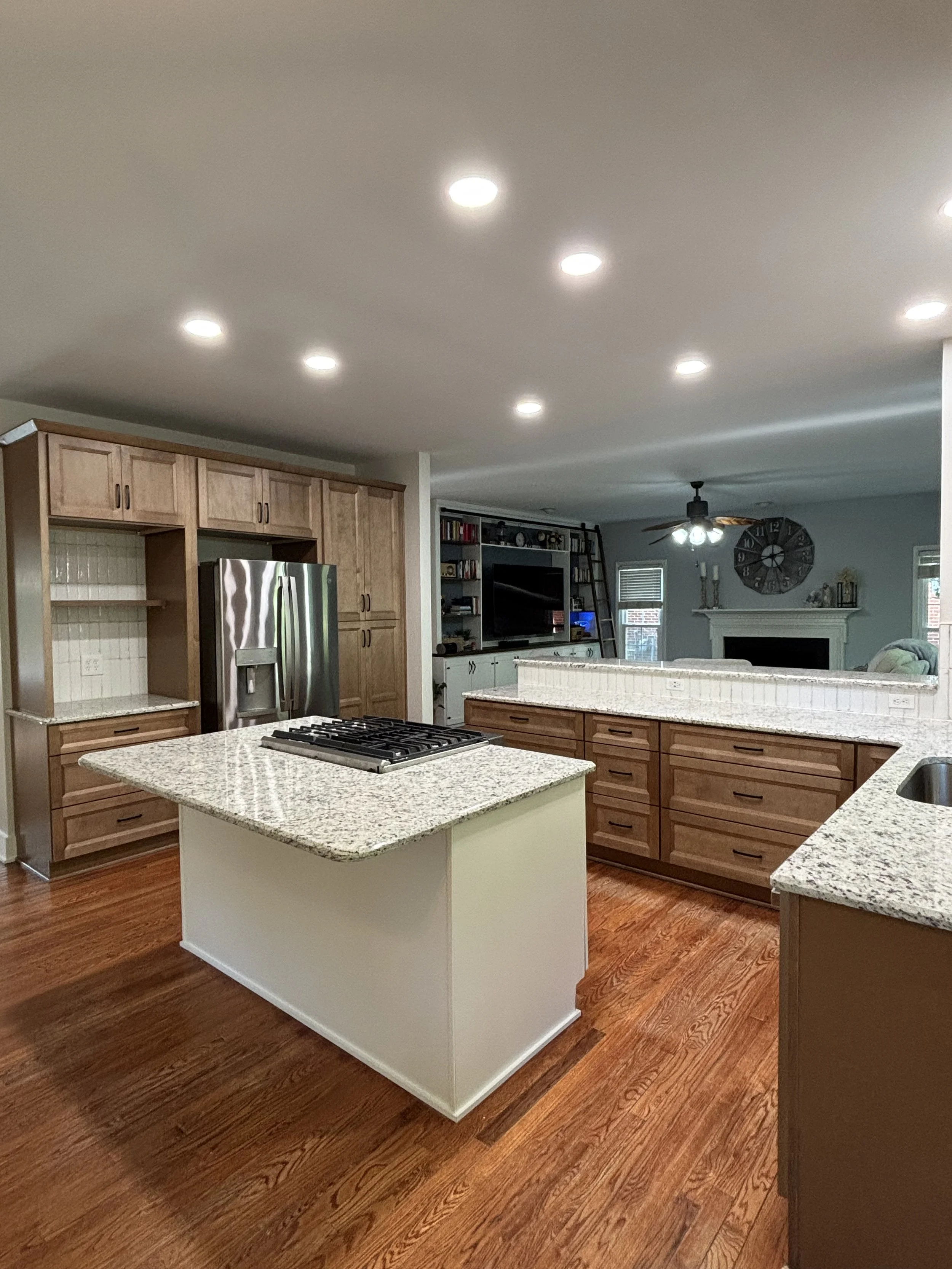 A modern kitchen with wooden cabinets, granite countertops, stainless steel refrigerator, island with a stove, hardwood floors, and a living room in the background with a fireplace, bookshelf, and clock.