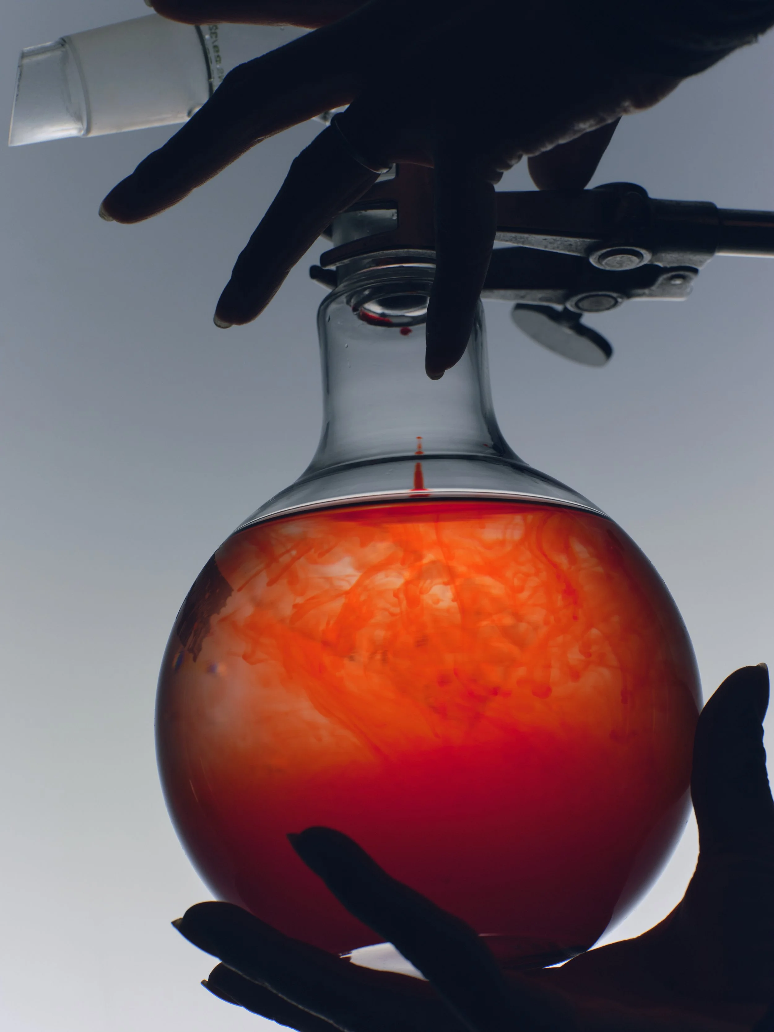 A scientist or researcher wearing black gloves handling a glass flask containing a red-orange liquid with swirling patterns, against a gray background.