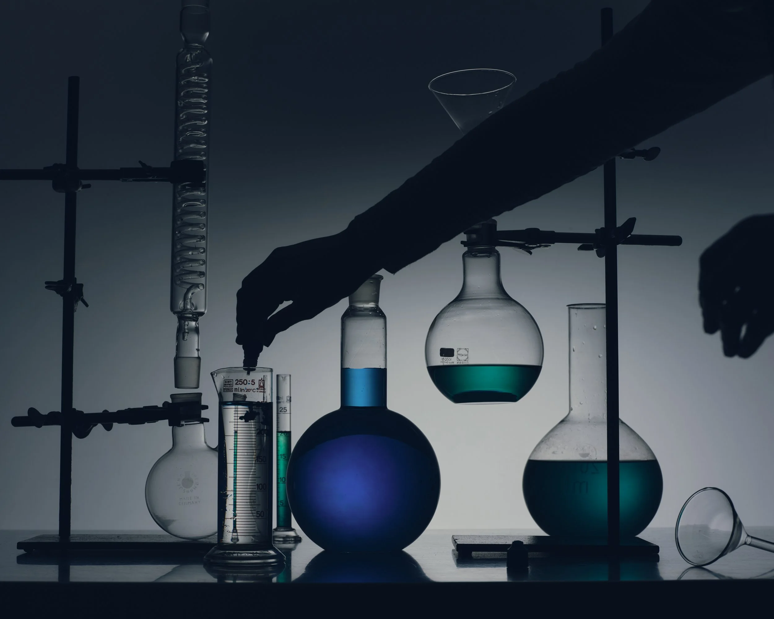 Silhouetted hands working with laboratory glassware and equipment, including flasks, beakers, and test tubes, with liquids of various colors.