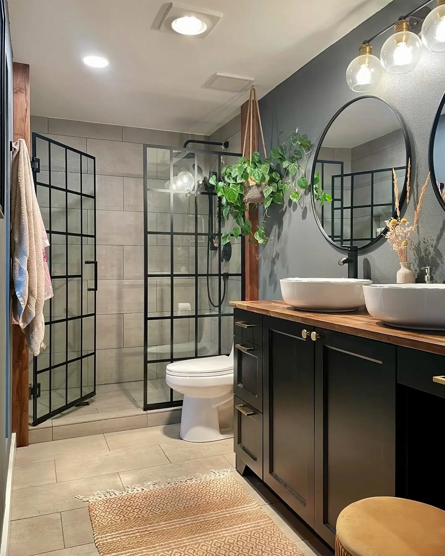 A modern bathroom with a walk-in shower enclosed by black-framed glass doors, a black vanity with twin vessel sinks, round mirrors, hanging plants, and decorative items.