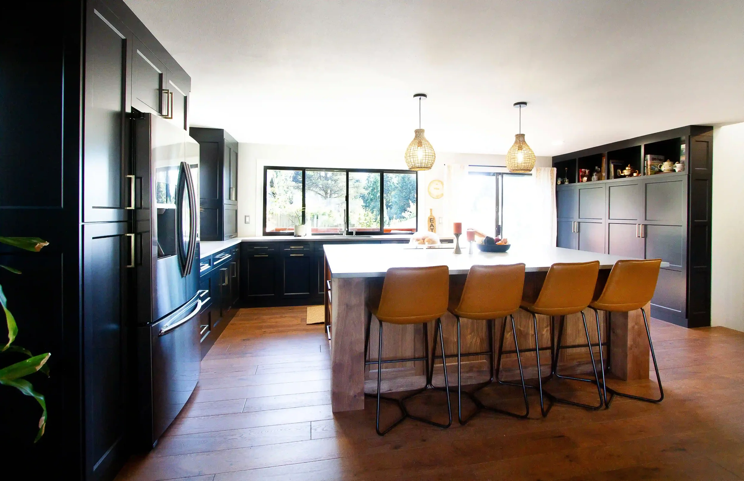 Modern kitchen with black cabinets, a large island with four tan barstools, wooden flooring, and pendant lights hanging above the island.