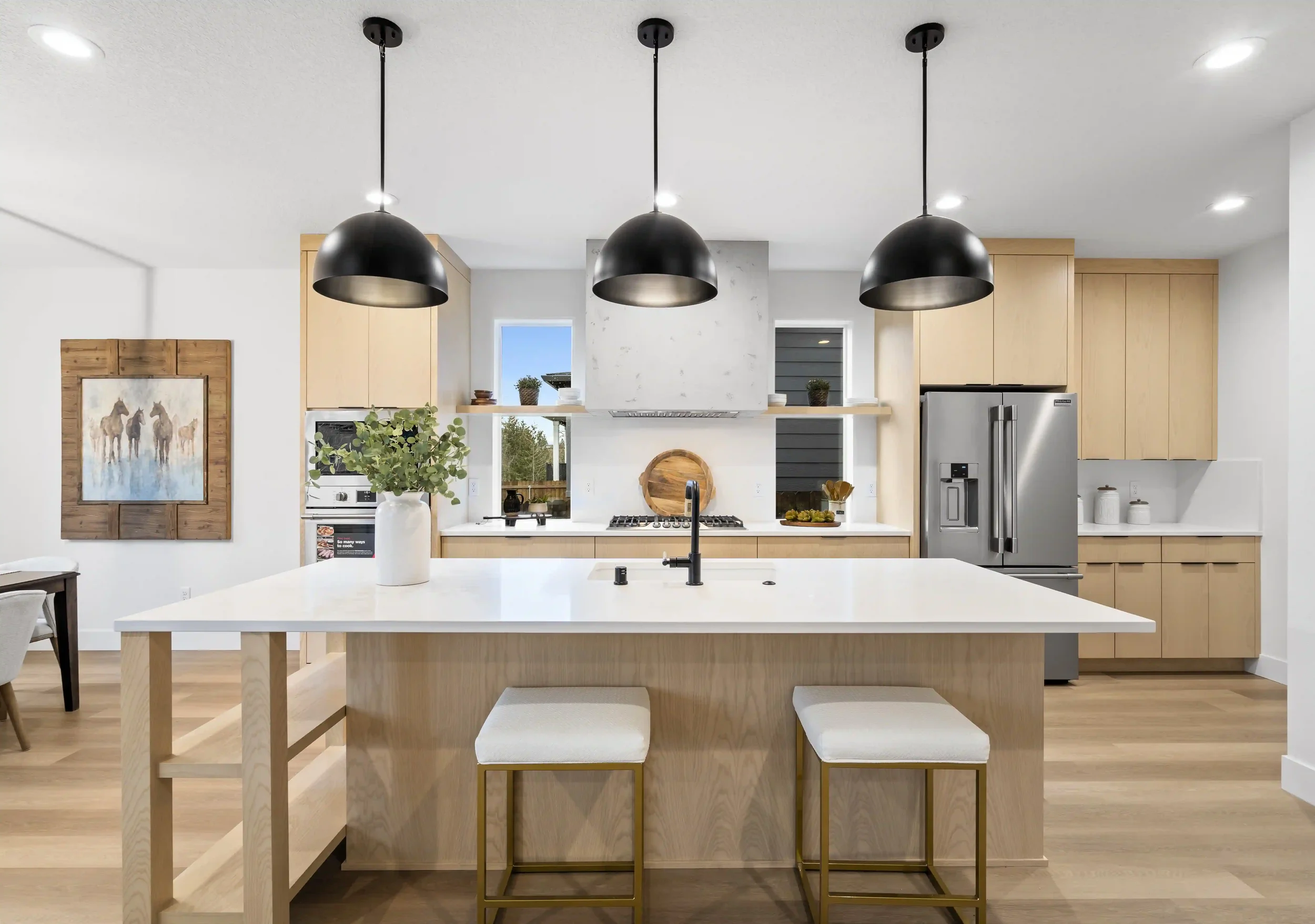 Modern kitchen with light wood cabinets, white countertops, black pendant lights, stainless steel refrigerator, and a large island with two stools.