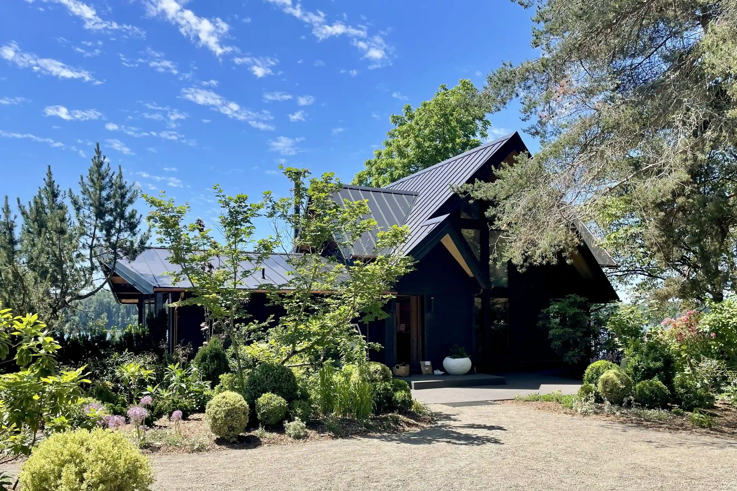 Modern house with dark exterior and metal roof, surrounded by lush greenery and trees, under a bright blue sky with scattered clouds.