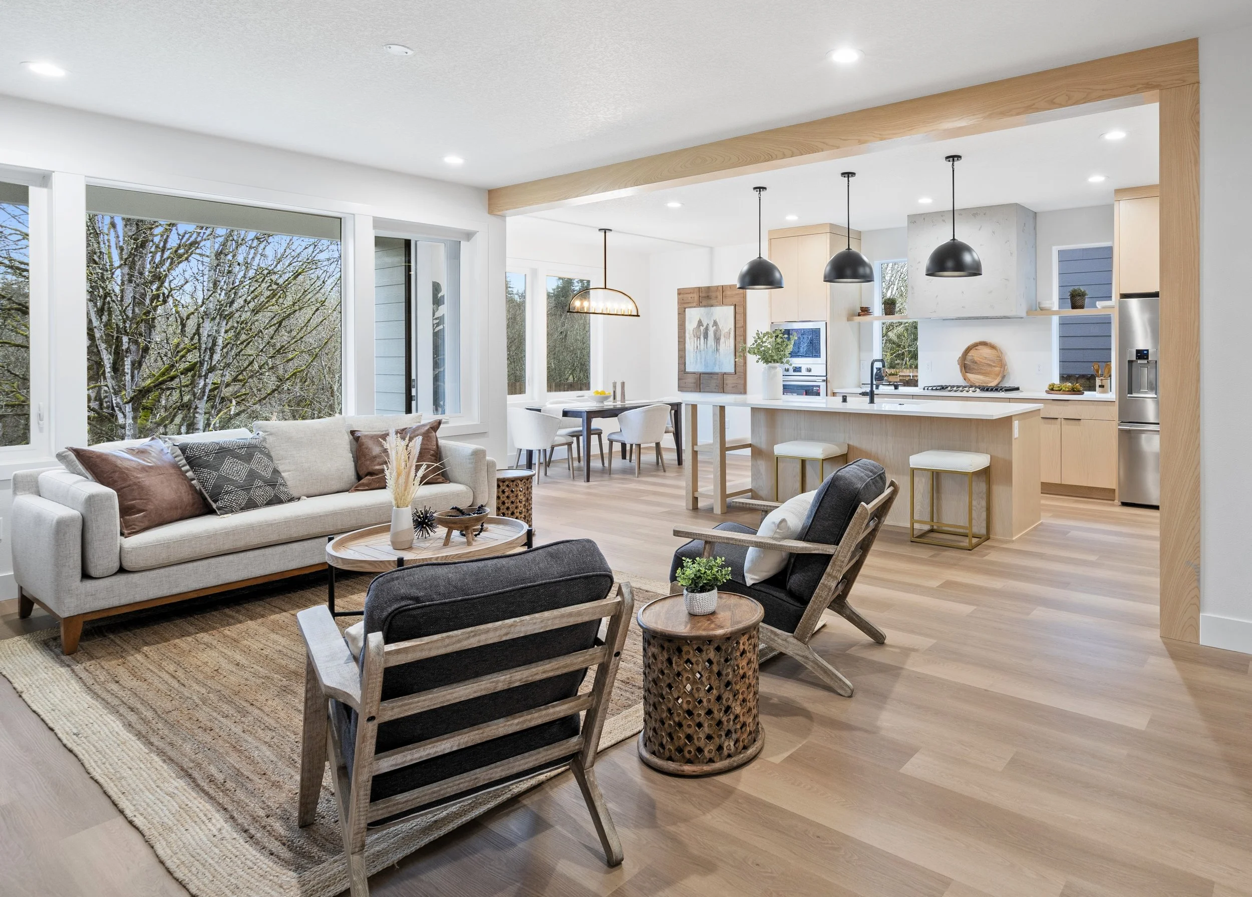 Open-concept living room and kitchen with large windows, light wood flooring, modern furniture, black pendant lights, and a view of trees outside.