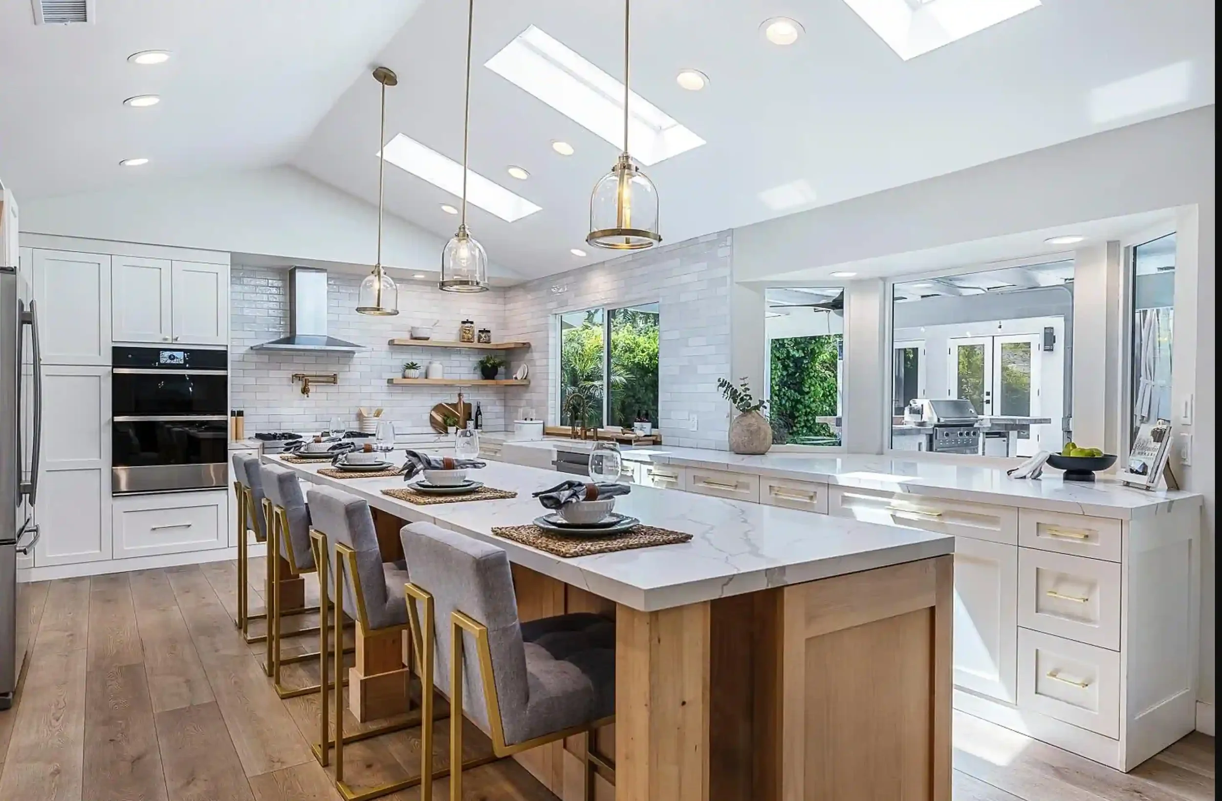 Modern white kitchen with a large island, gray chairs with gold frames, pendant lights, open shelving, appliances, and large windows showing greenery outside.