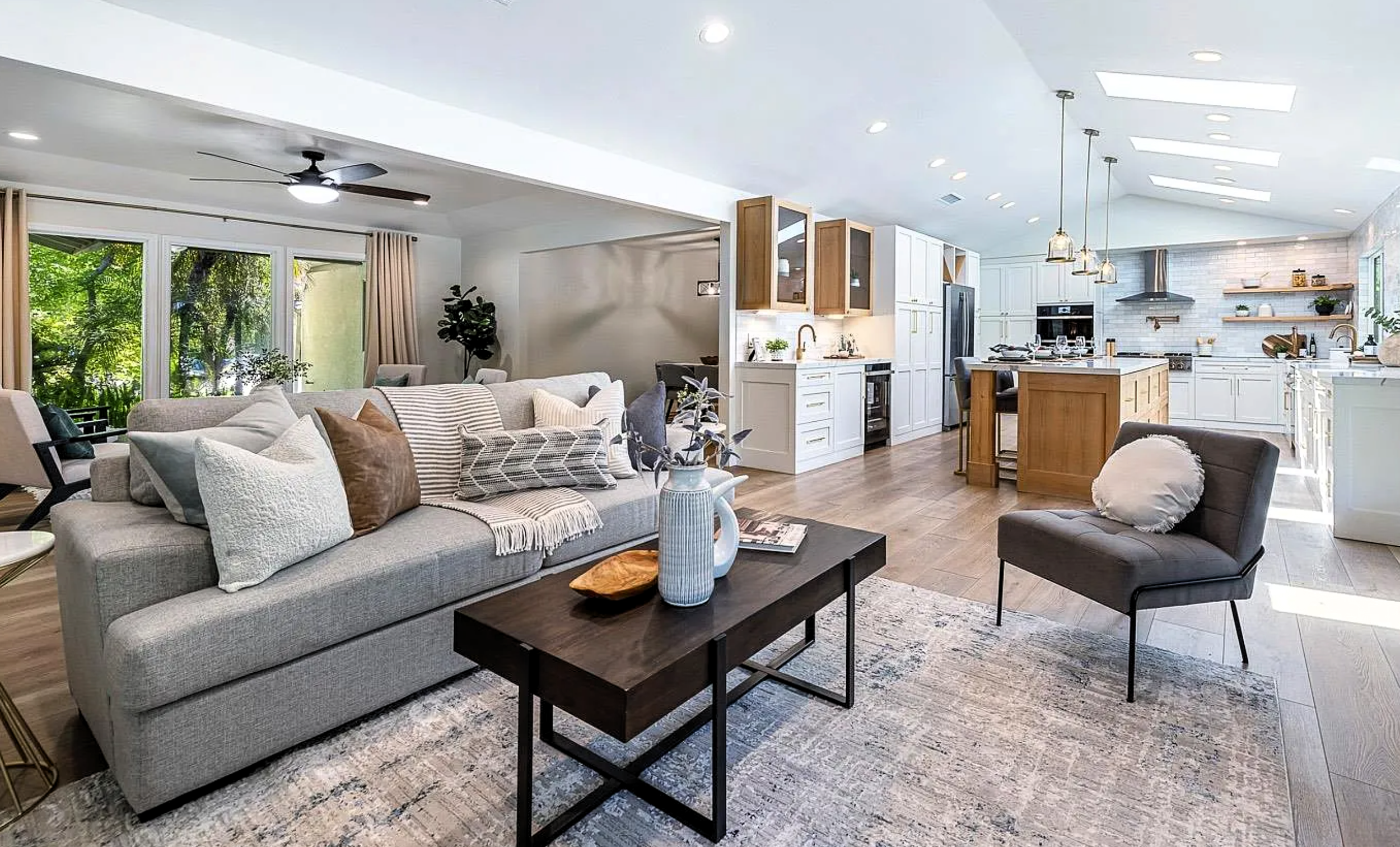 Open concept living room and kitchen with neutral tones, modern furnishings, and large windows showing greenery outside.