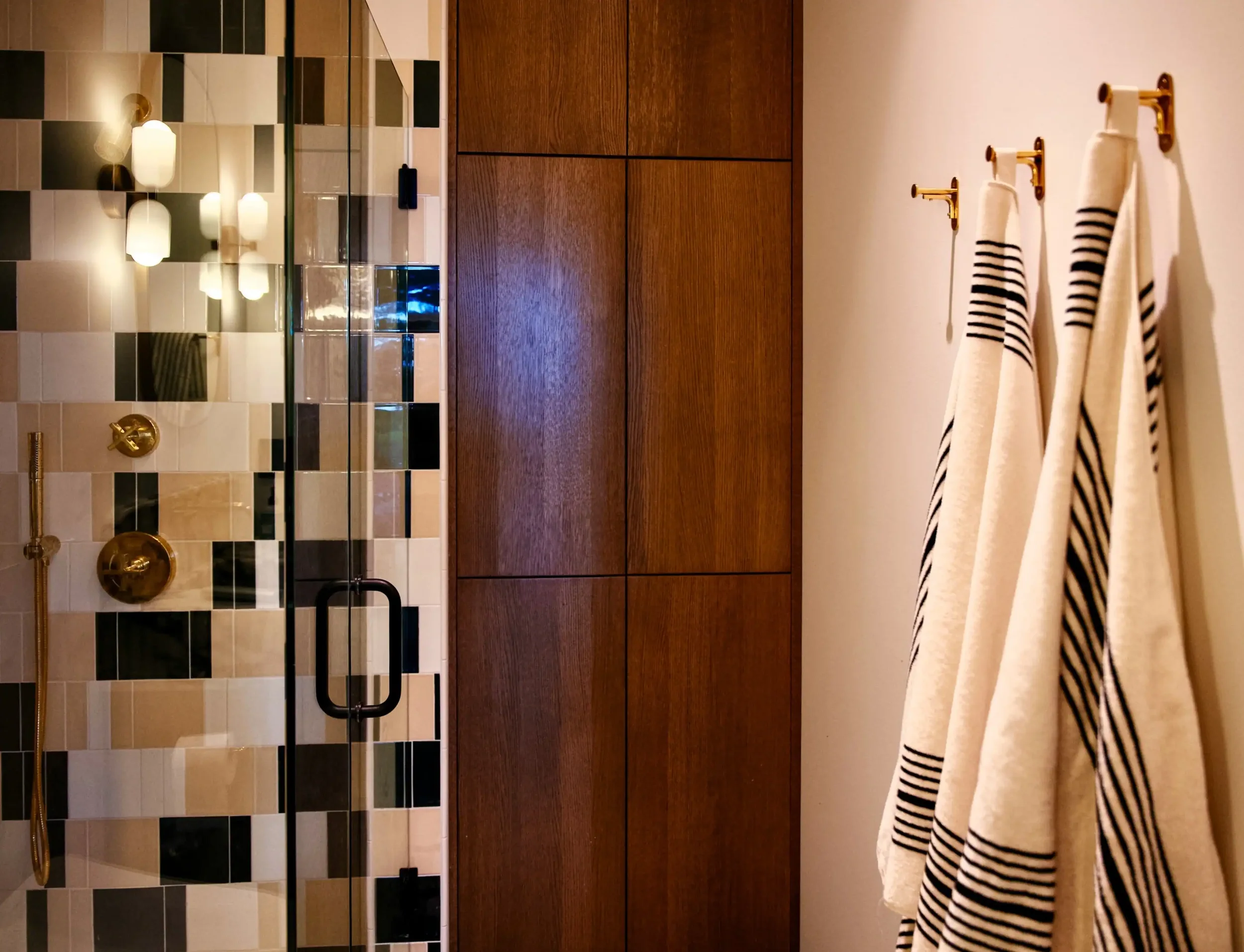 A modern bathroom with a glass-enclosed shower featuring black, beige, and cream tiles, and a gold showerhead. There are three hanging striped towels on wall hooks to the right, and wooden cabinetry on the wall behind the shower.