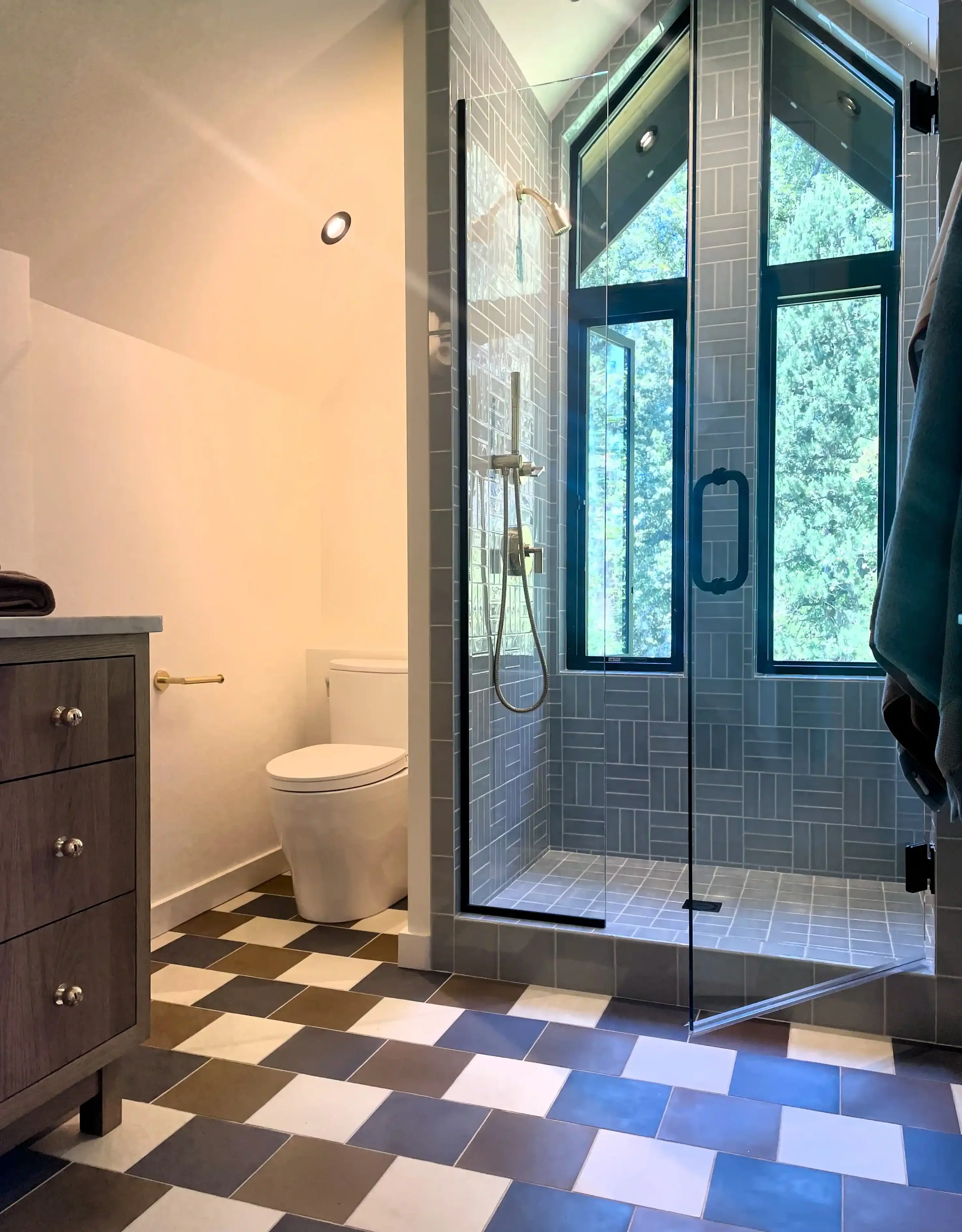 Modern bathroom with a glass-enclosed shower featuring blue tile and large windows, a toilet, and a dark wood vanity with drawers, with a checkered black, white, and beige floor.