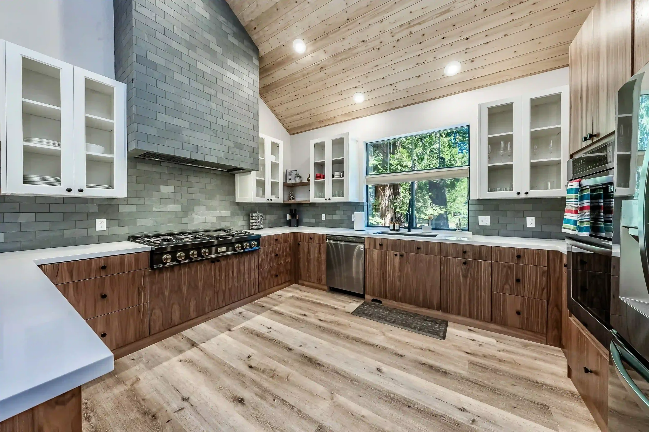 Modern kitchen with wooden lower cabinets, white upper cabinets with glass doors, gray subway tile backsplash, stainless steel appliances, a large window, wood-paneled ceiling, and light wood flooring.