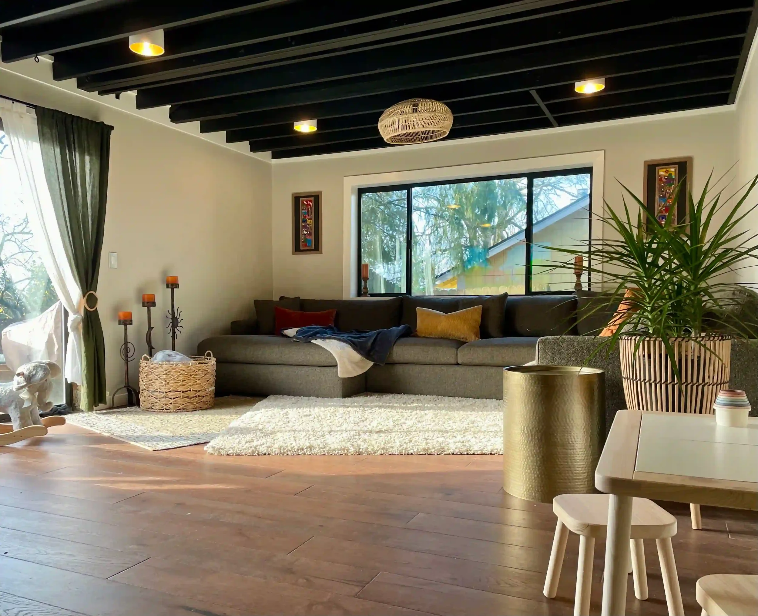 Living room with large sectional sofa, potted plant, white shaggy rug, wooden flooring, and large window letting in natural light.