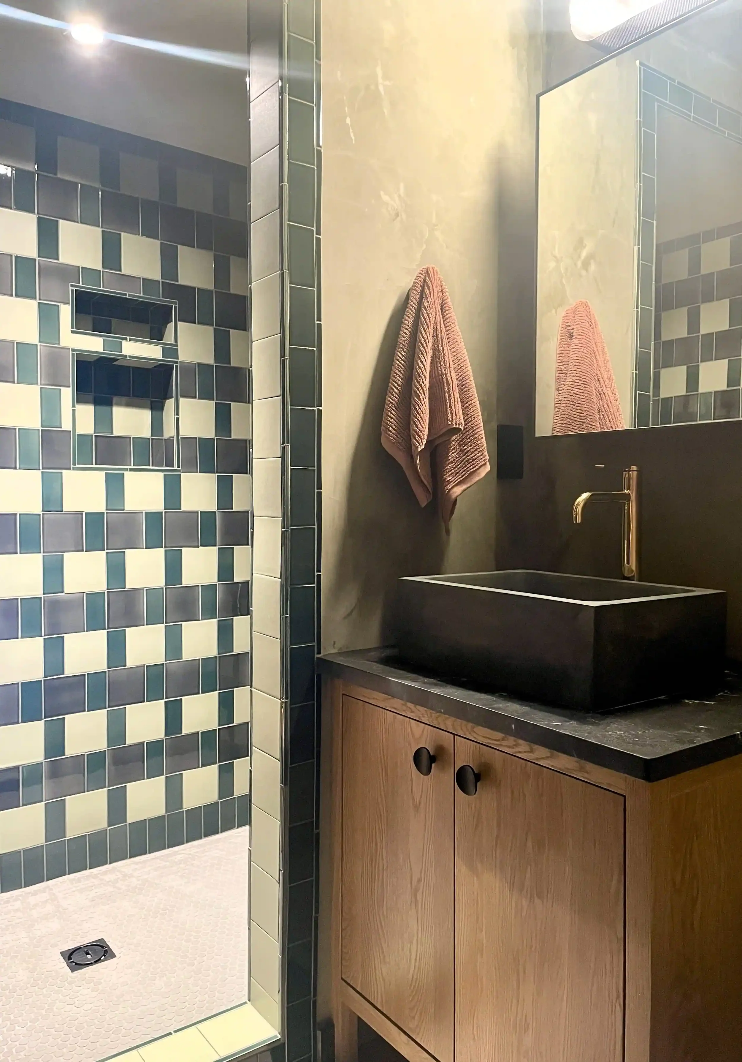 Modern bathroom with a wood cabinet sink, pink towel, decorative black vessel sink, and a tiled shower area with blue and cream tiles.