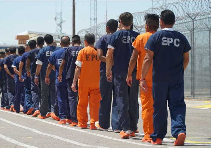 Detainees being escorted across a courtyard.  Blue uniforms indicate no criminal history.