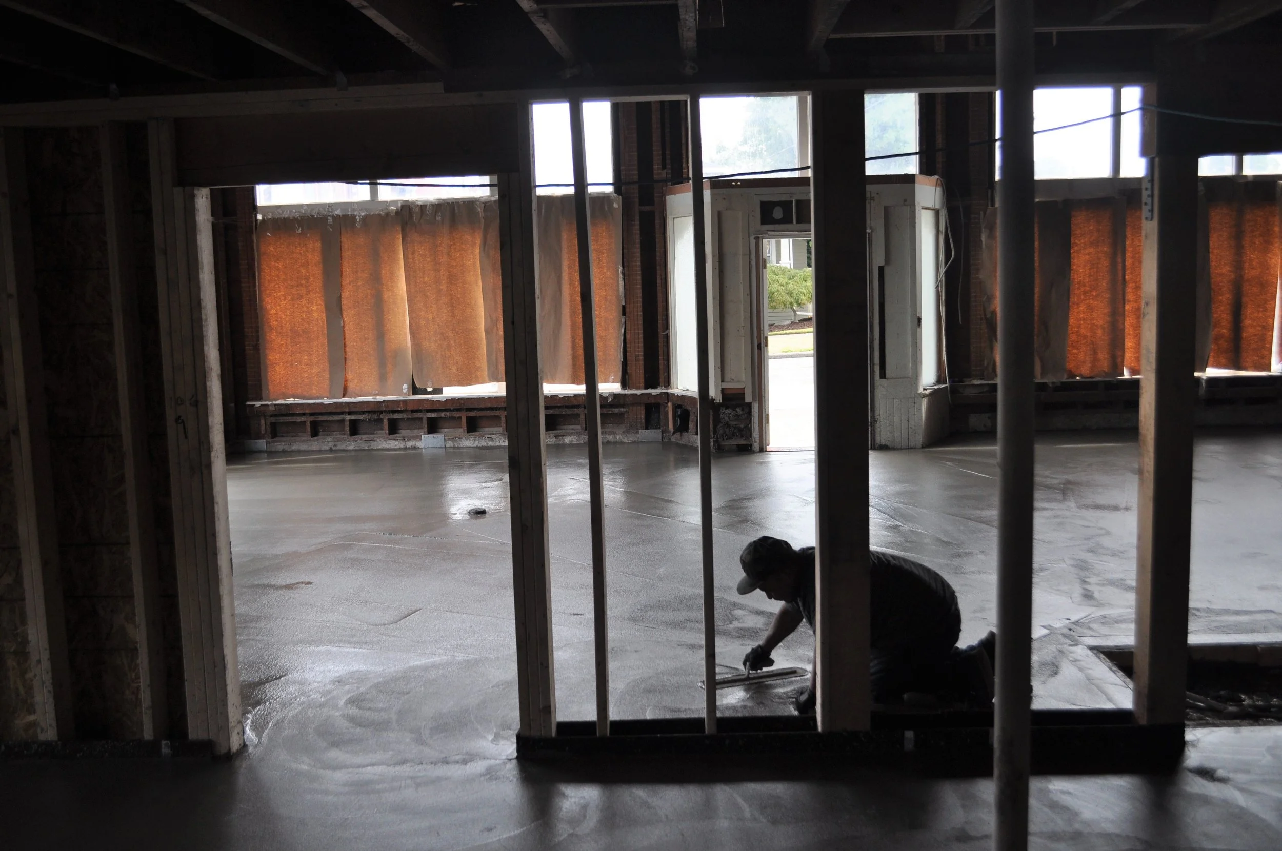 A person kneeling on a newly poured concrete floor inside an unfinished building, with framing and windows installed and some curtains hanging.