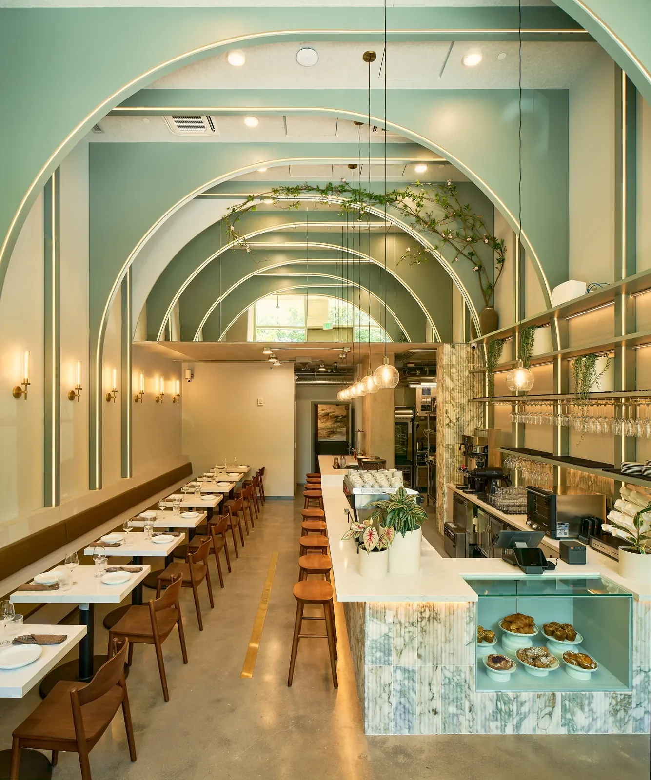 Modern restaurant interior with a long bar counter, white marble finishes, and green decor accents. The ceiling has curved arches with lighting, and the space features a mix of seating and greenery with decorative plants.