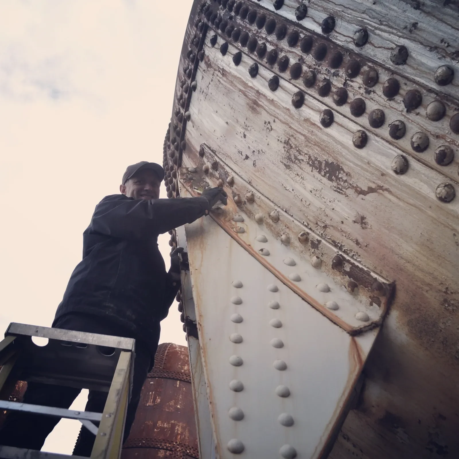 A man on a ladder inspecting and working on the rivets and structure of a large industrial tank or vessel outdoors.