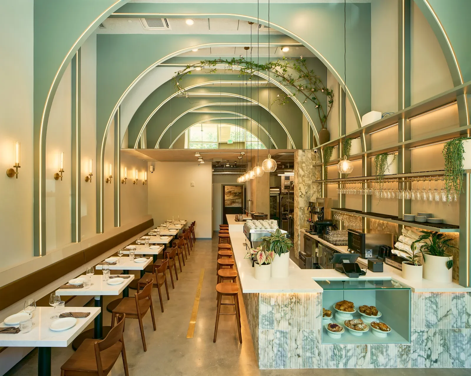 Modern restaurant interior with a light-colored marble bar, hanging globe lights, a row of dining tables with chairs, and decorative plants and arches