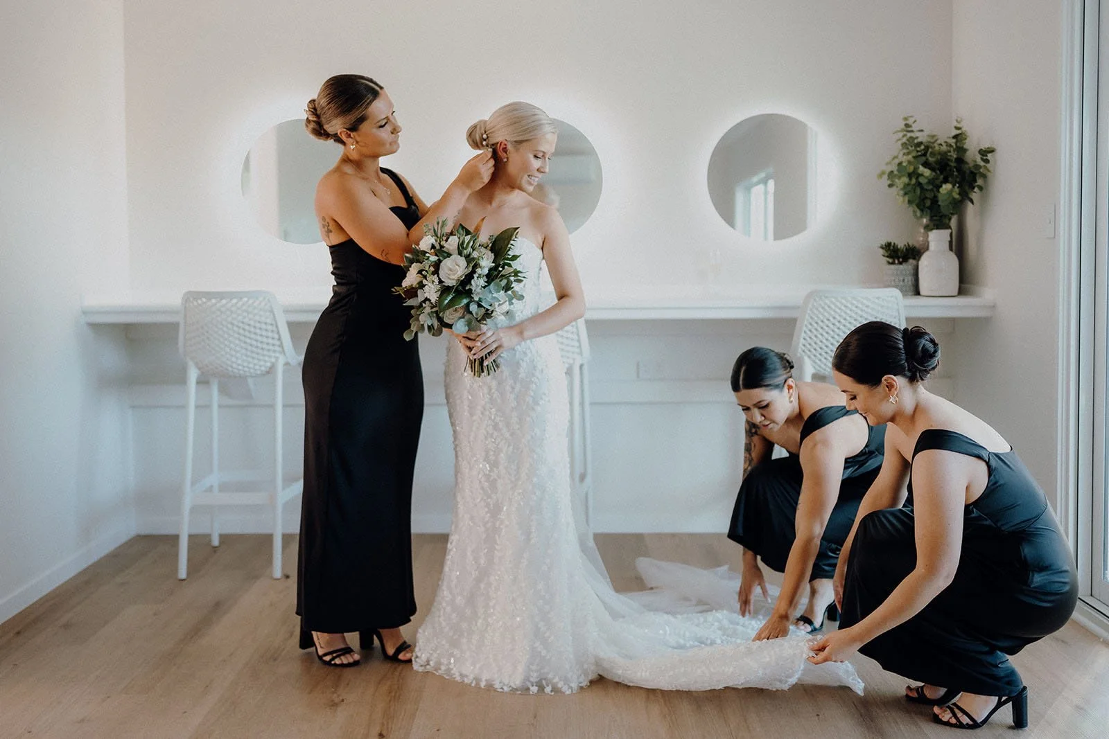Cowbell Creek Wedding Venue - A bride in a white lace wedding gown is assisted by her bridesmaids in black dresses as they prepare her gown
