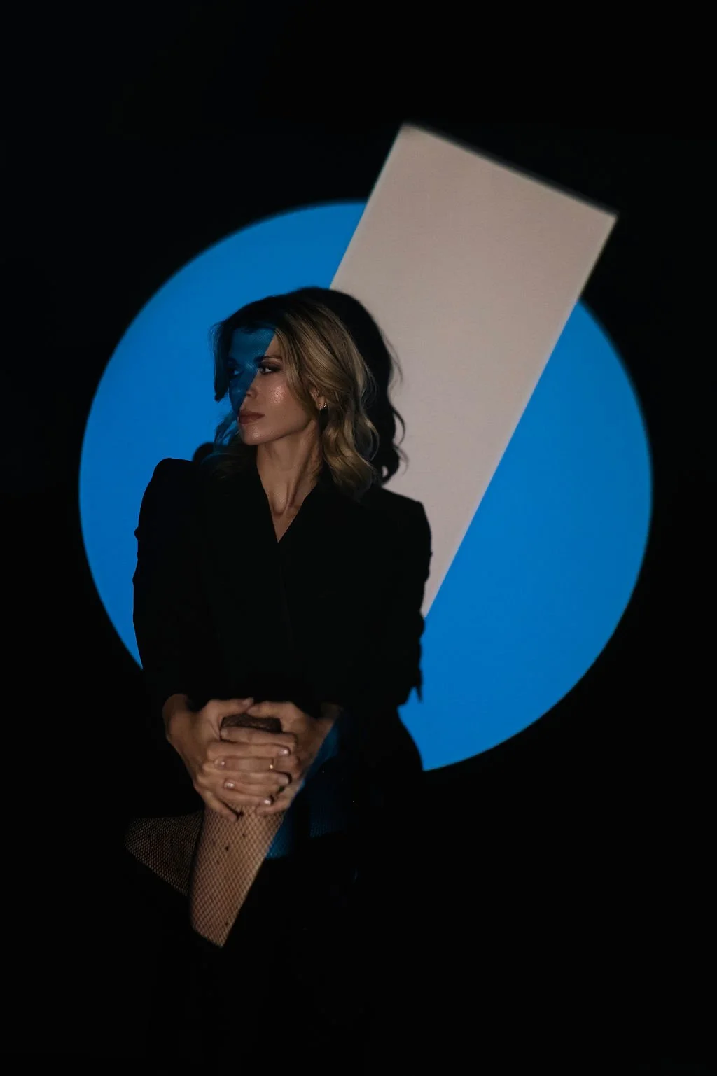 A woman with wavy blonde hair and dark makeup, wearing a black blazer with crossed arms, standing in front of a background with a blue circle and a vertical white rectangle.
