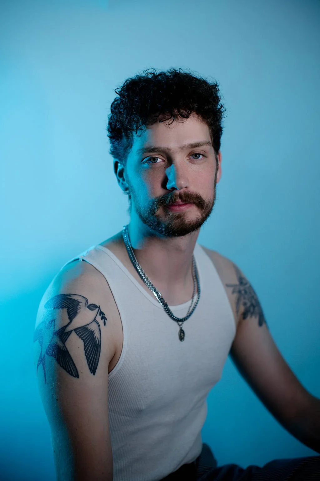 Portrait of a man with curly hair and a beard, wearing a white tank top, silver necklace, seated against a blue background.