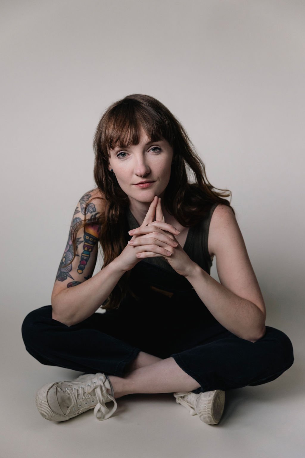 Young woman with long brown hair, sitting cross-legged on the floor with her hands under her chin, wearing a black sleeveless top, black pants, and white sneakers, against a plain light-colored background.