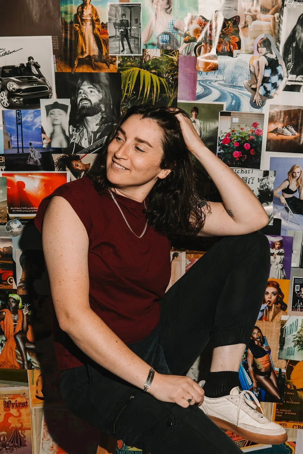 Young woman with dark hair, wearing a red shirt, sitting in front of a collage of various posters and photographs, smiling gently with her hand resting on her head.