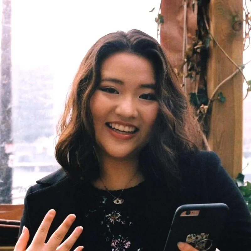 Young woman with wavy brown hair, smiling, holding a smartphone, sitting in a cozy indoor setting with a wooden post and ivy in the background.