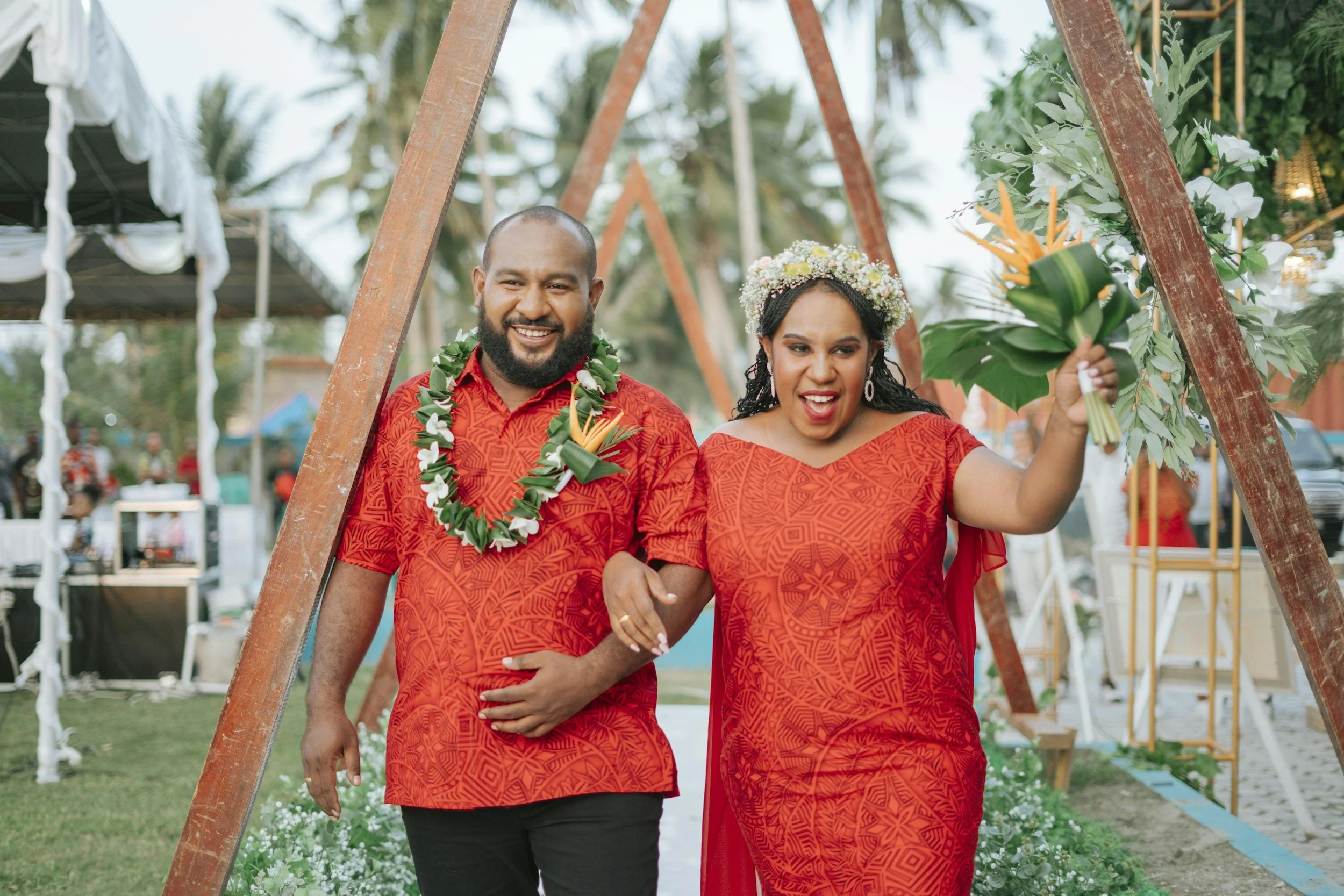 A smiling couple walking outdoors during a celebration, both wearing matching red traditional clothing, with the woman holding a bouquet and wearing a flower crown, and the man wearing a floral lei.