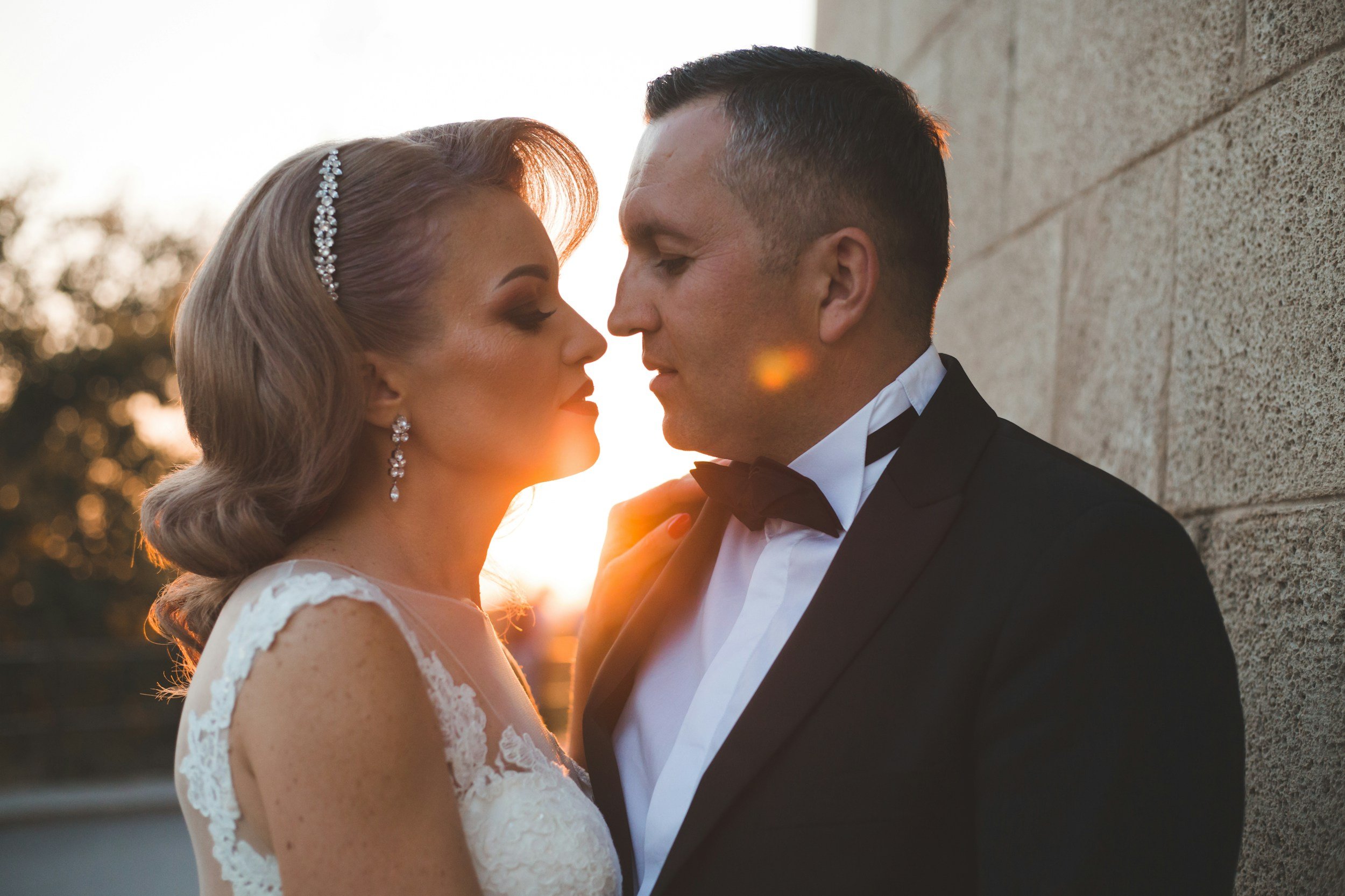 A bride and groom face each other closely outdoors at sunset, with the bride in a lace wedding dress and jewelry, and the groom in a tuxedo, with a wall behind them.