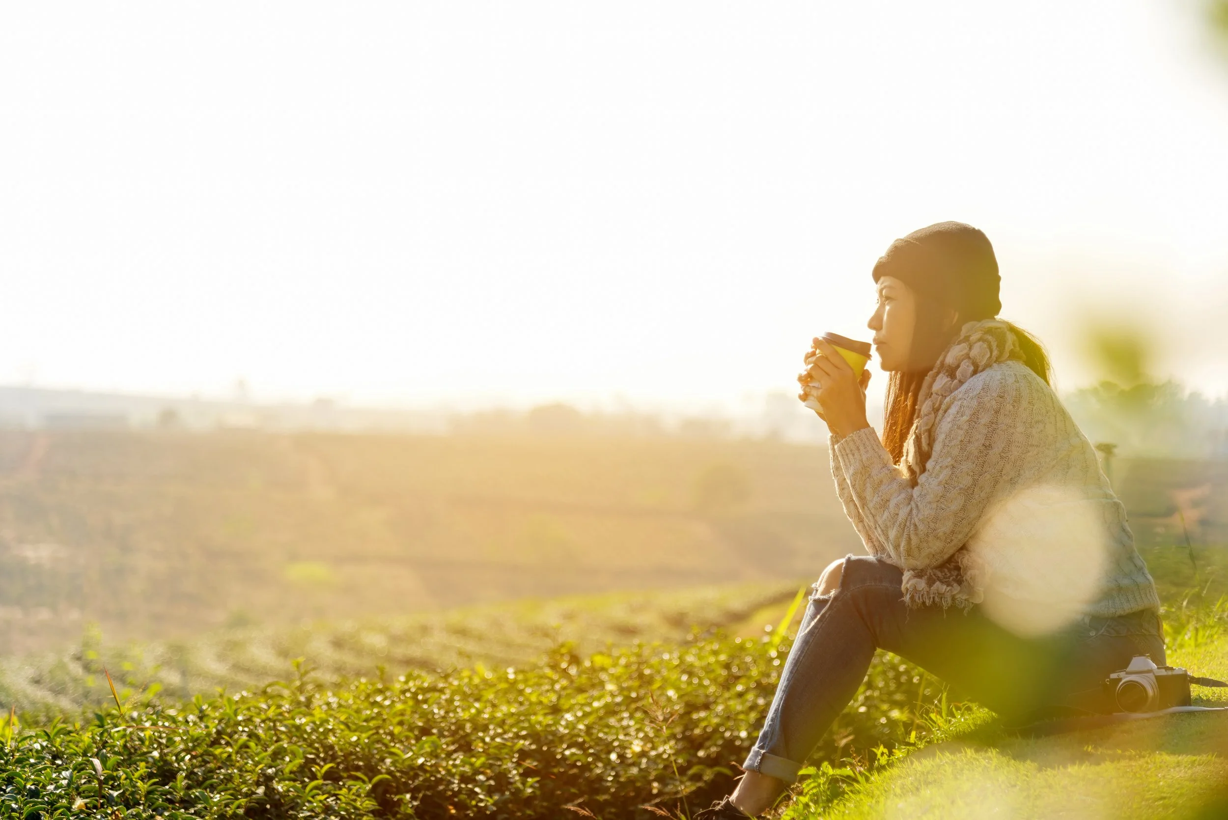 A woman sitting outdoors on grass in a field, holding a yellow mug and enjoying a warm beverage at sunset.