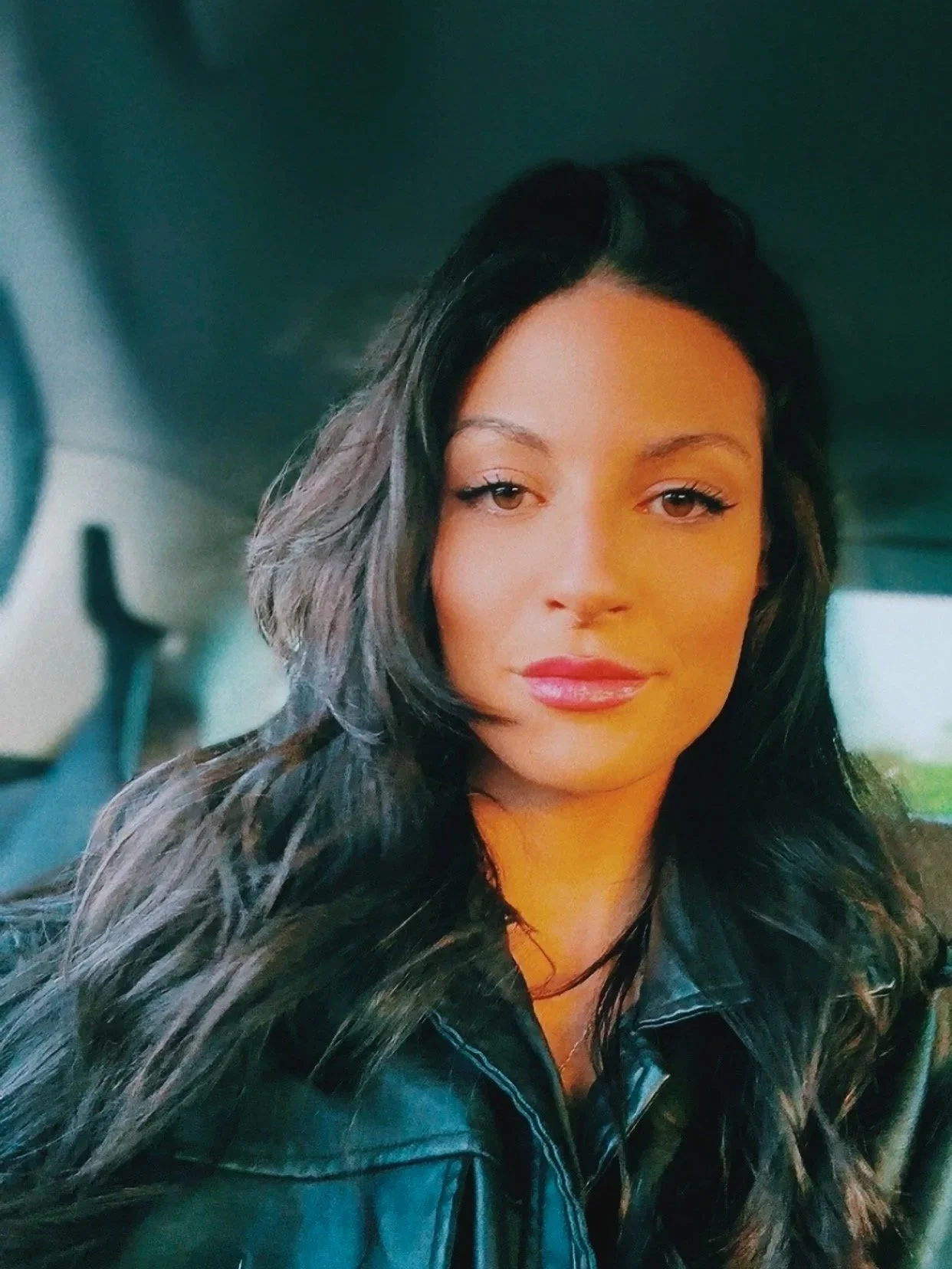 A woman with long wavy dark hair and makeup, wearing a black leather jacket, sitting in a vehicle.