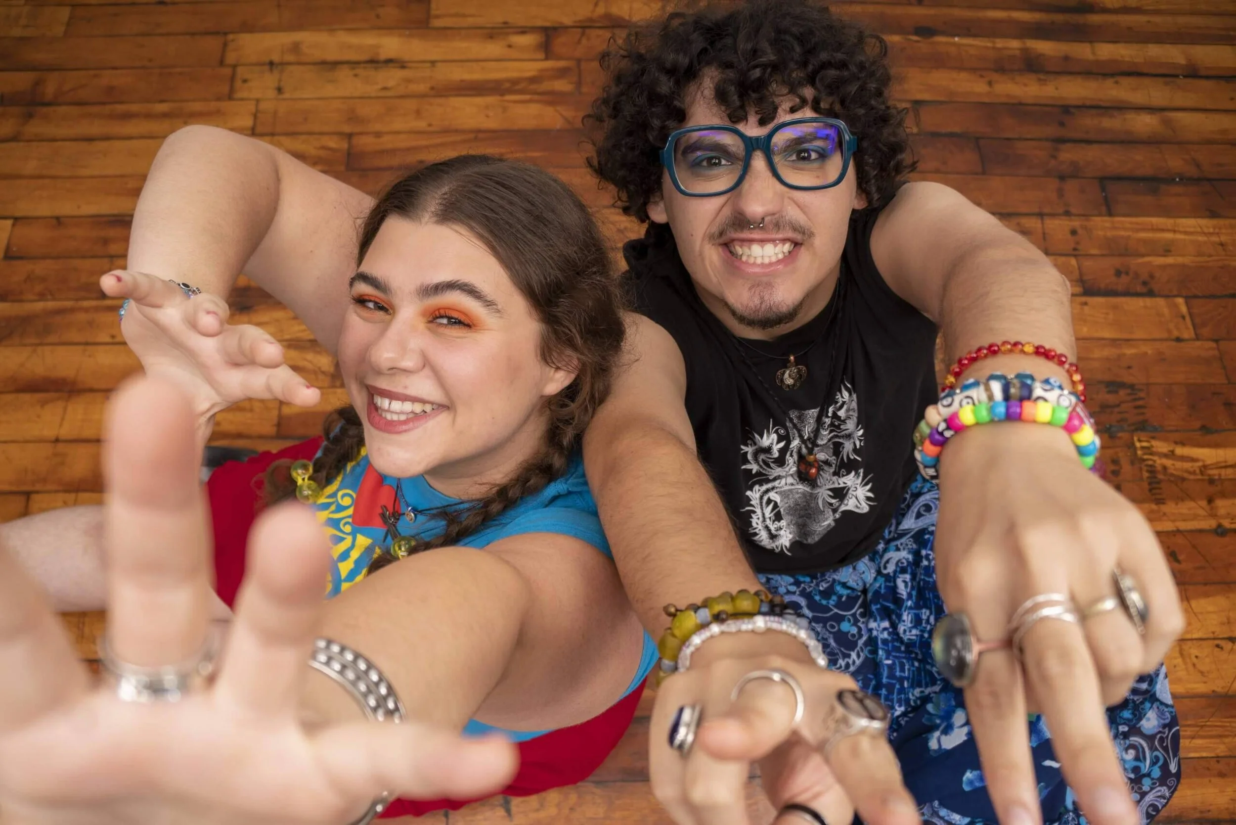 Two young adults with colorful jewelry and glasses, smiling and reaching towards the camera, sitting on a wooden floor.
