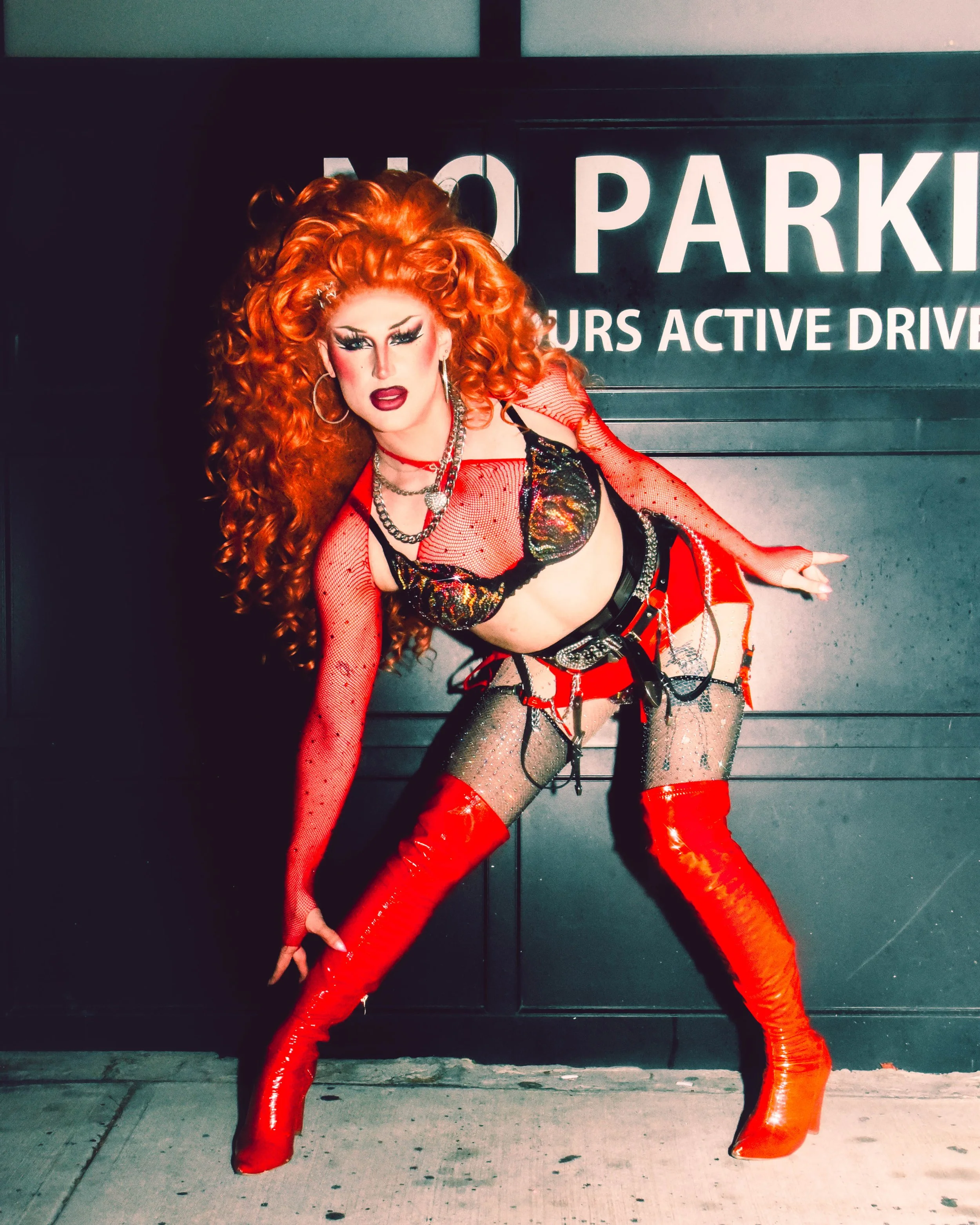 A drag queen with voluminous curly red hair, bold makeup, wearing a revealing black and red outfit with thigh-high red boots, posing in front of a parking garage sign.
